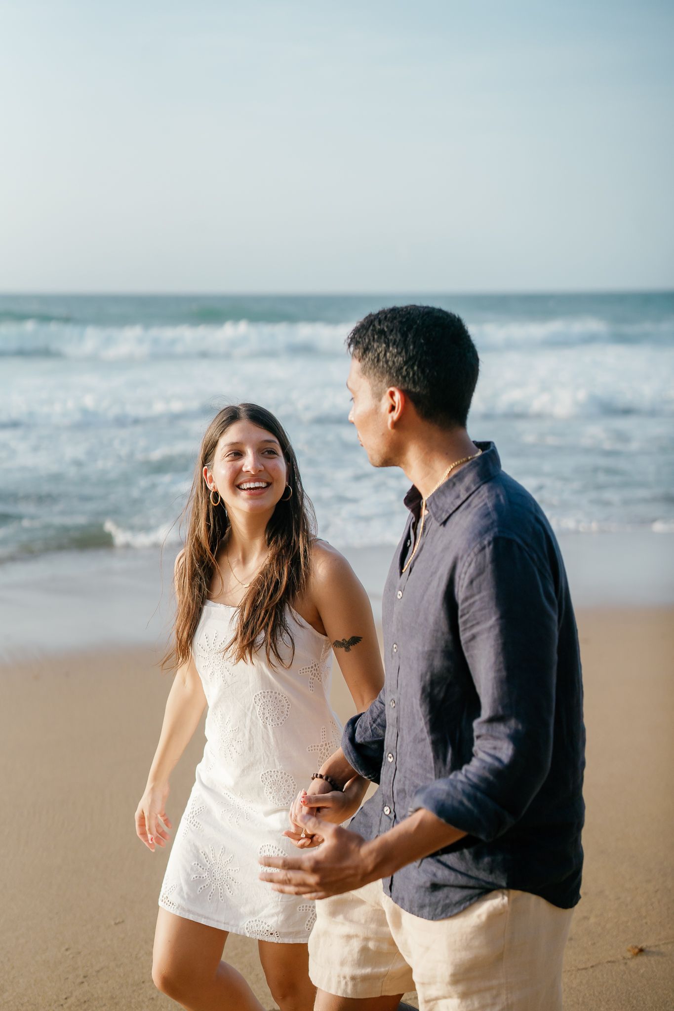 A couple shares joyful moments together while walking along a sandy beach with ocean waves in the background.
