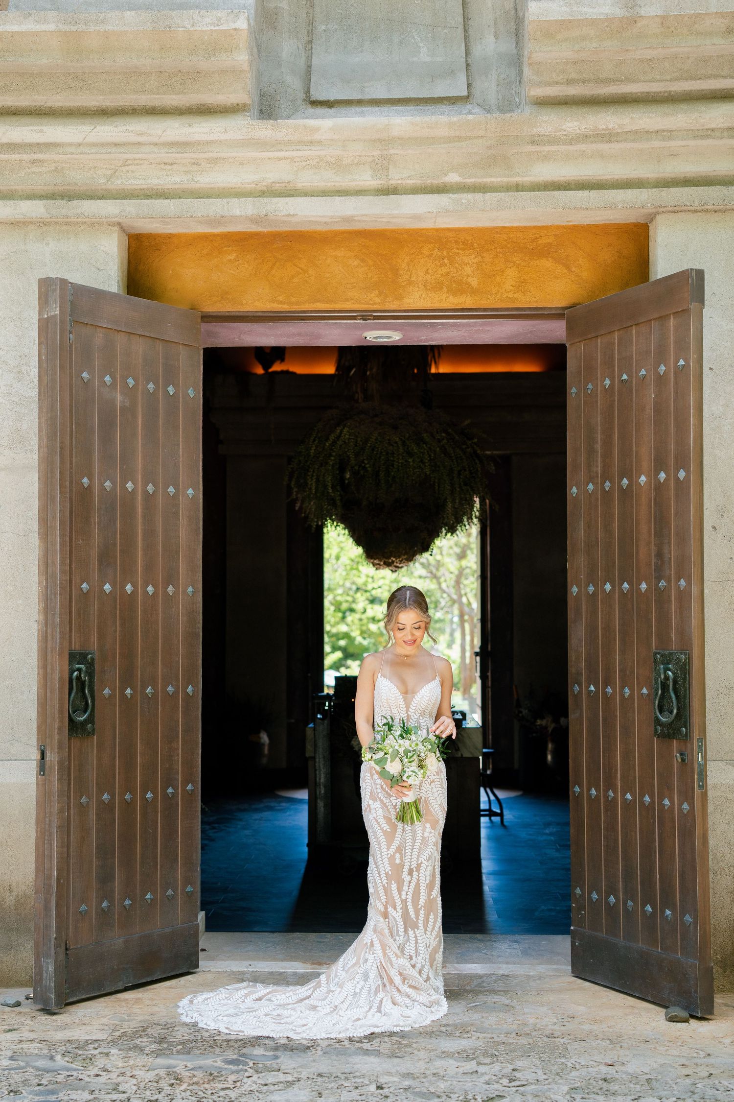 A figure in a flowing white lace wedding dress stands elegantly framed by large wooden doorways at an ornate venue.