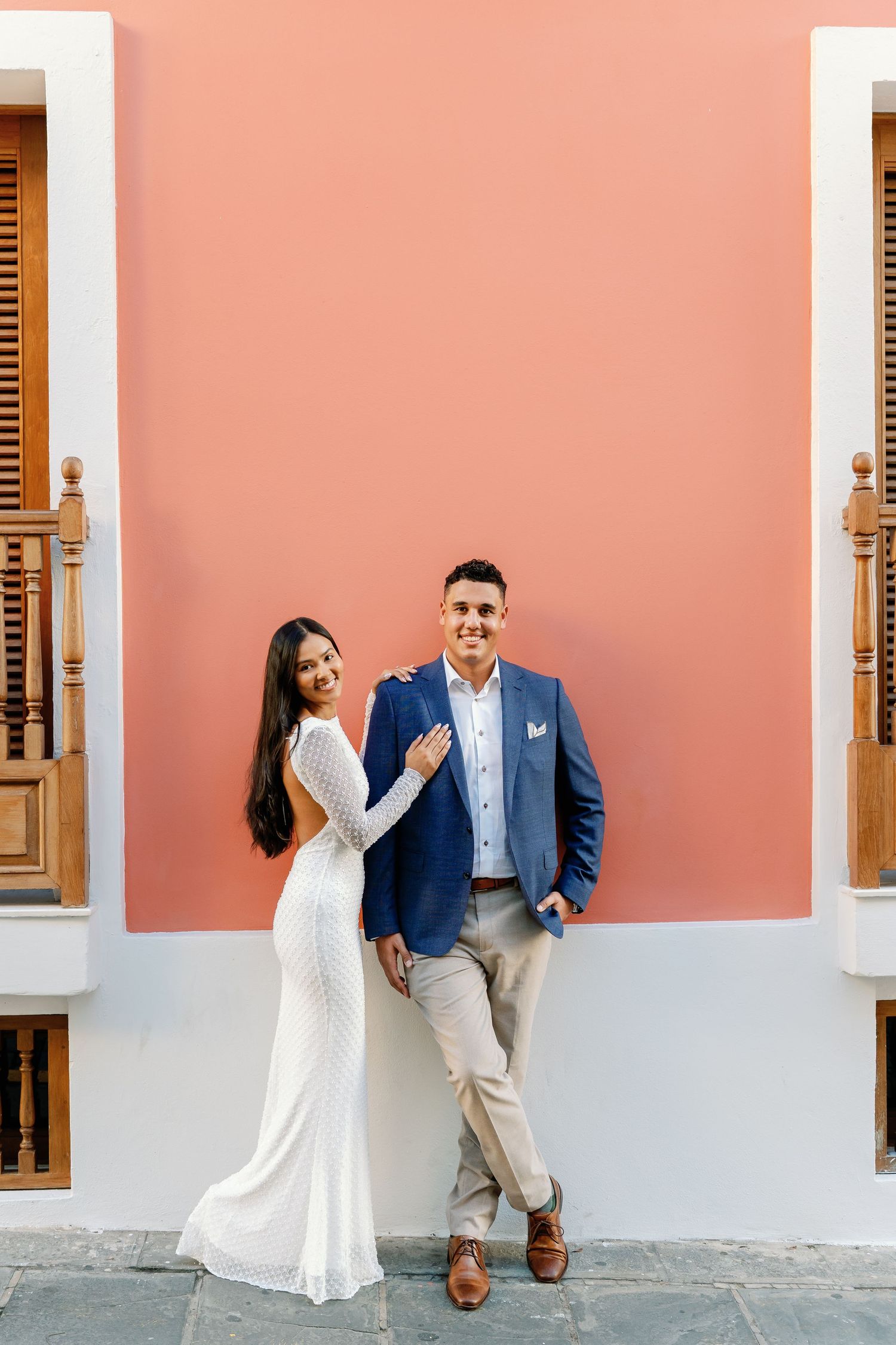 Couple in wedding attire posing against a coral pink wall, bride in white gown, groom in blue blazer.