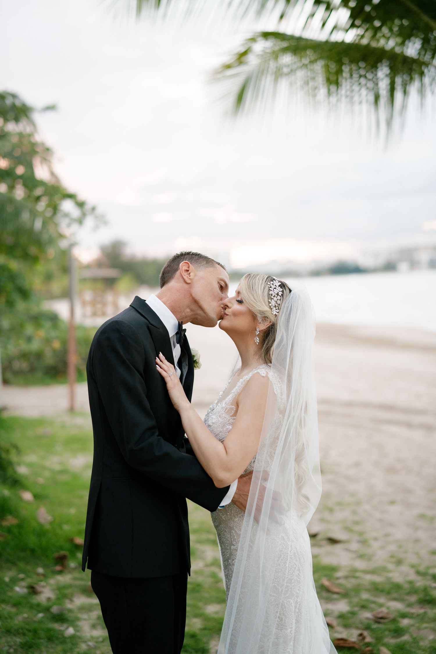 Bride and groom share a romantic beach kiss, she wears a lace gown with veil, he wears a black tuxedo.