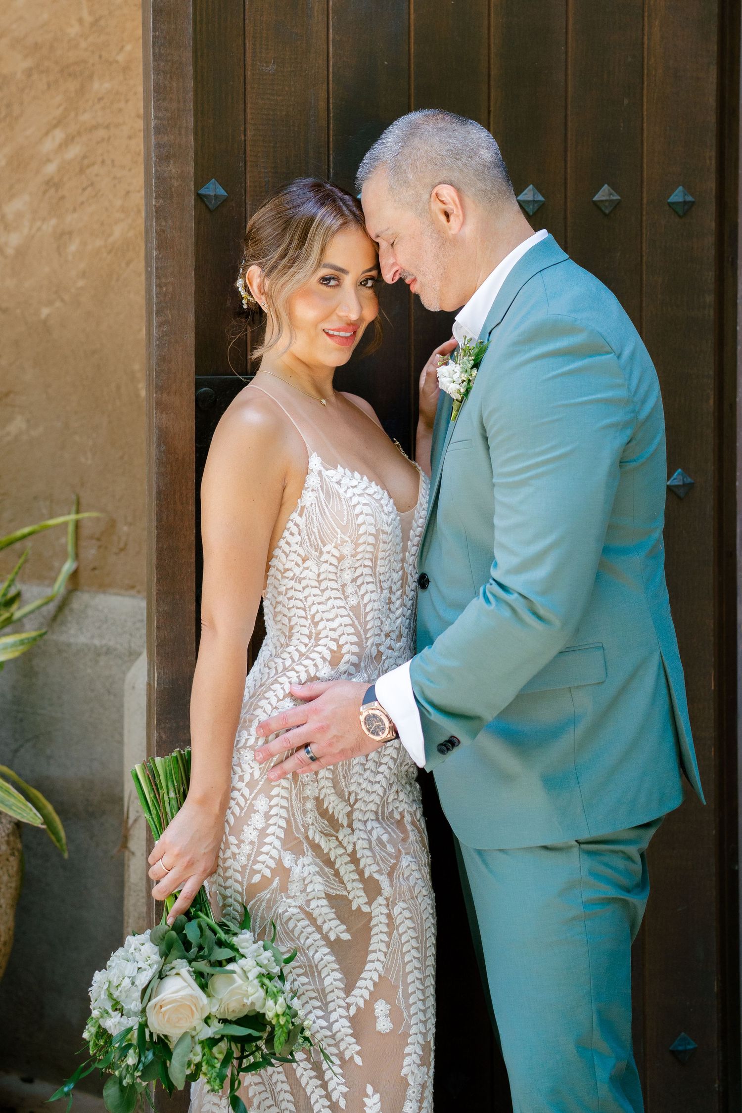 A couple embraces in formal wedding attire, with the bride holding a white and green bouquet against a rustic door.