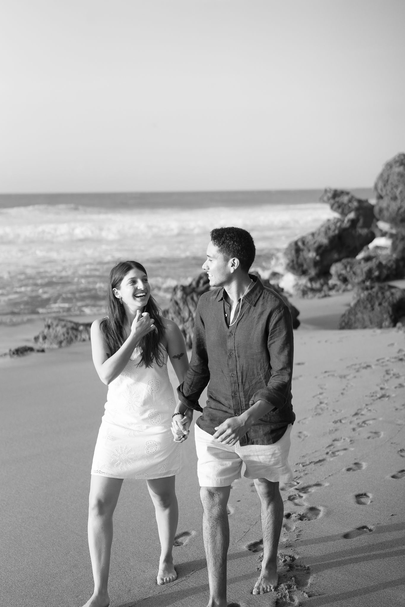 A couple walks playfully along a scenic beach with waves and rocks in black and white.