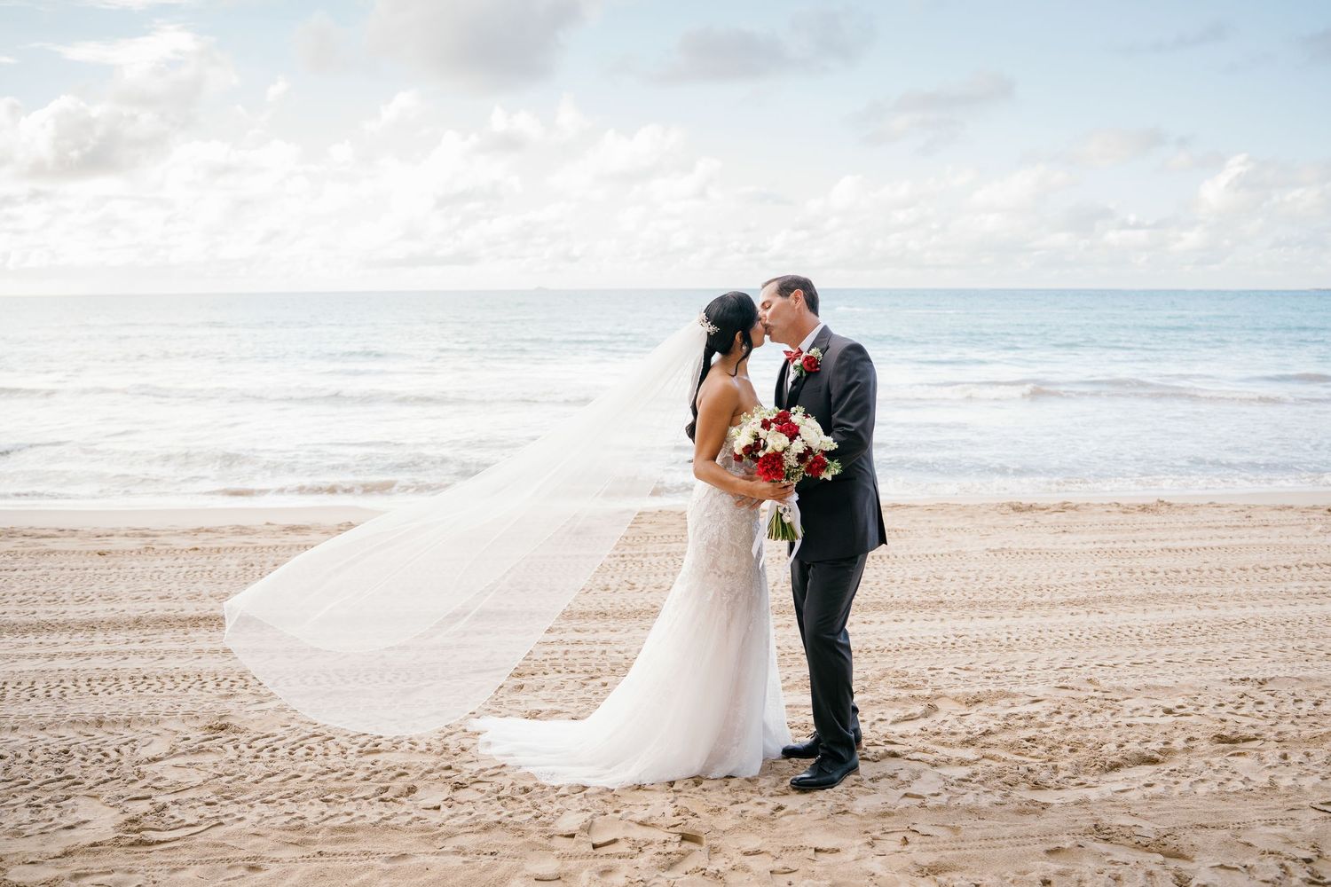 Bride and groom share a kiss on a sandy beach, her veil flowing in the wind, holding a red bouquet.