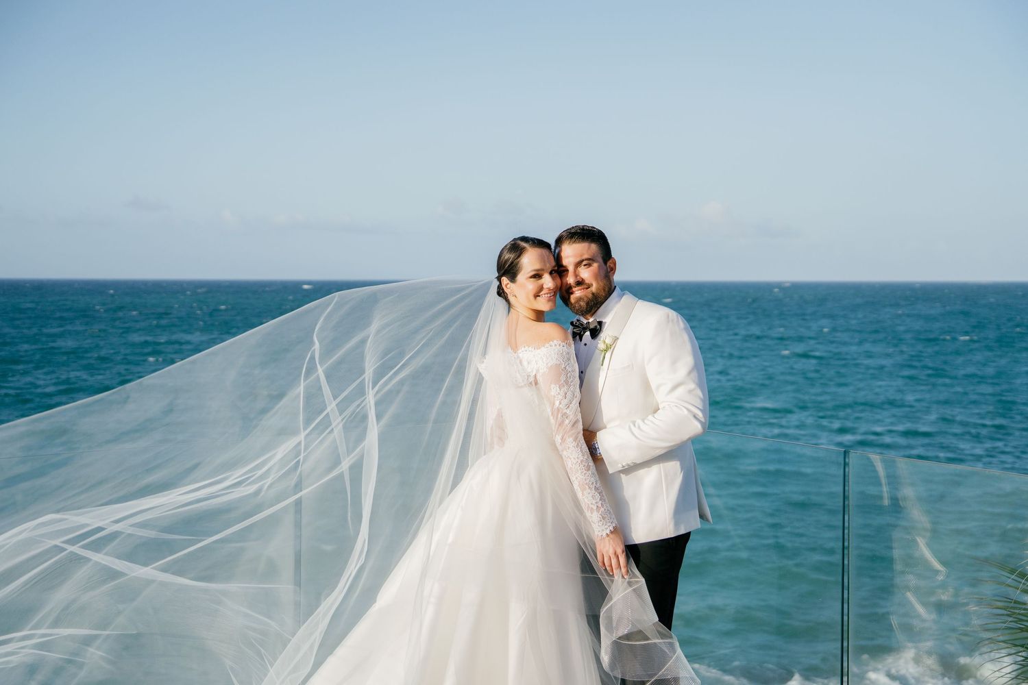 Bride and groom pose on oceanfront terrace, her veil flowing in the breeze, wearing white gown and white tuxedo.