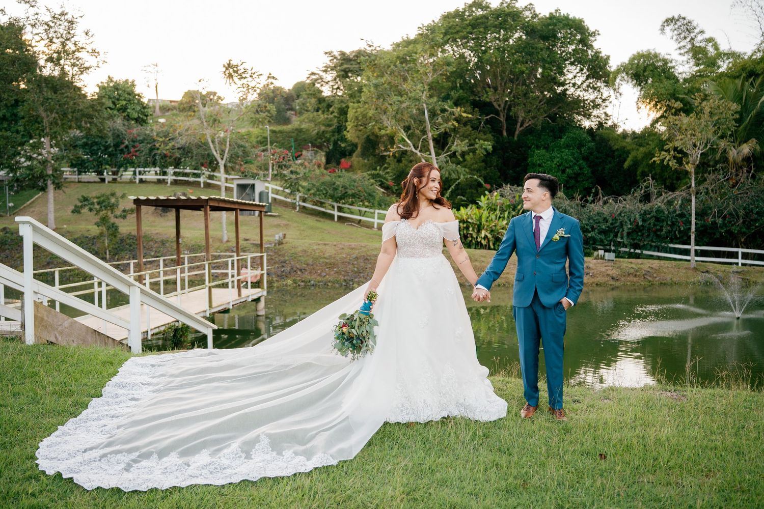 Bride in white gown with long train holds hands with groom in blue suit walking through lush garden near pond.