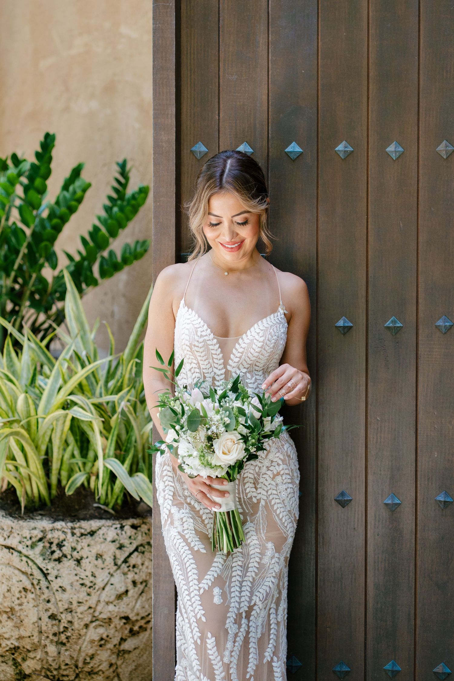 An elegant lace wedding dress with delicate beading is shown in multiple angles against a rustic wooden wall and greenery.