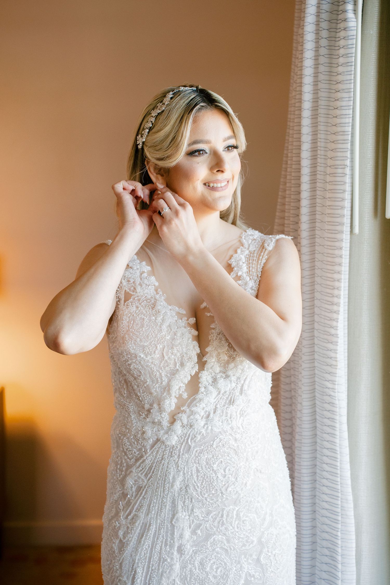 Bride in lace wedding gown adjusting earring near window with soft warm lighting.