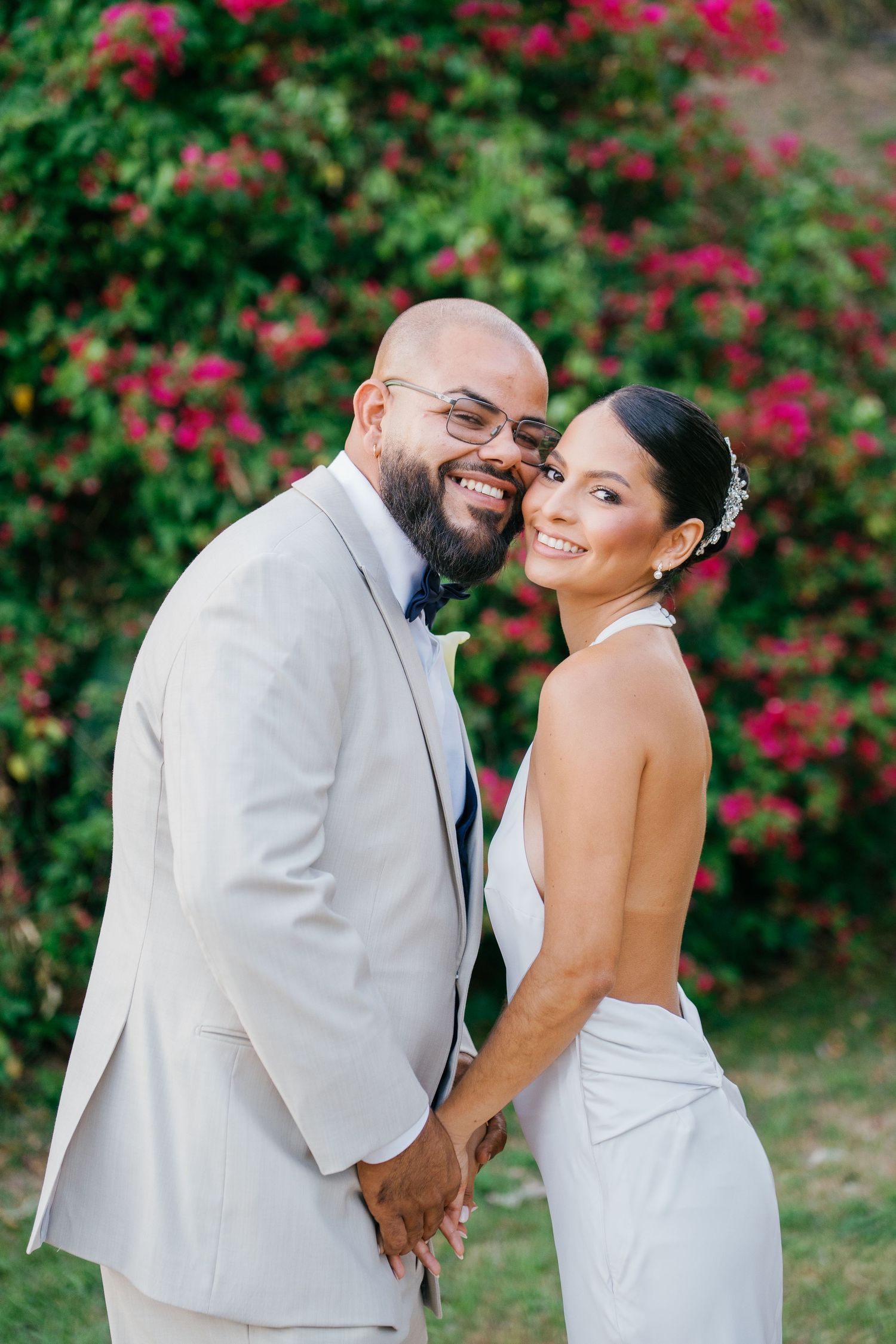Smiling bride and groom pose together in front of vibrant pink flowers, groom in beige suit, bride in white halter gown.