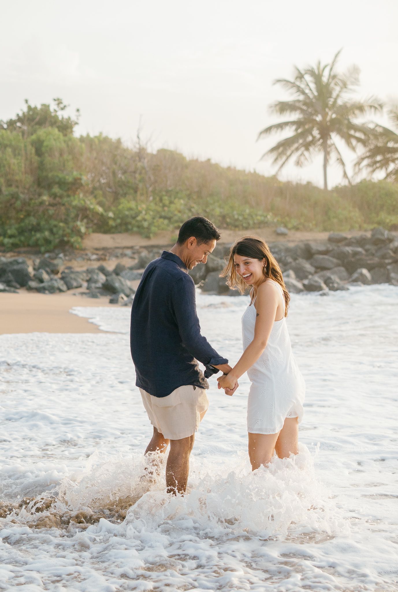 A couple splashes and plays together in the ocean waves at sunset on a tropical beach with palm trees.