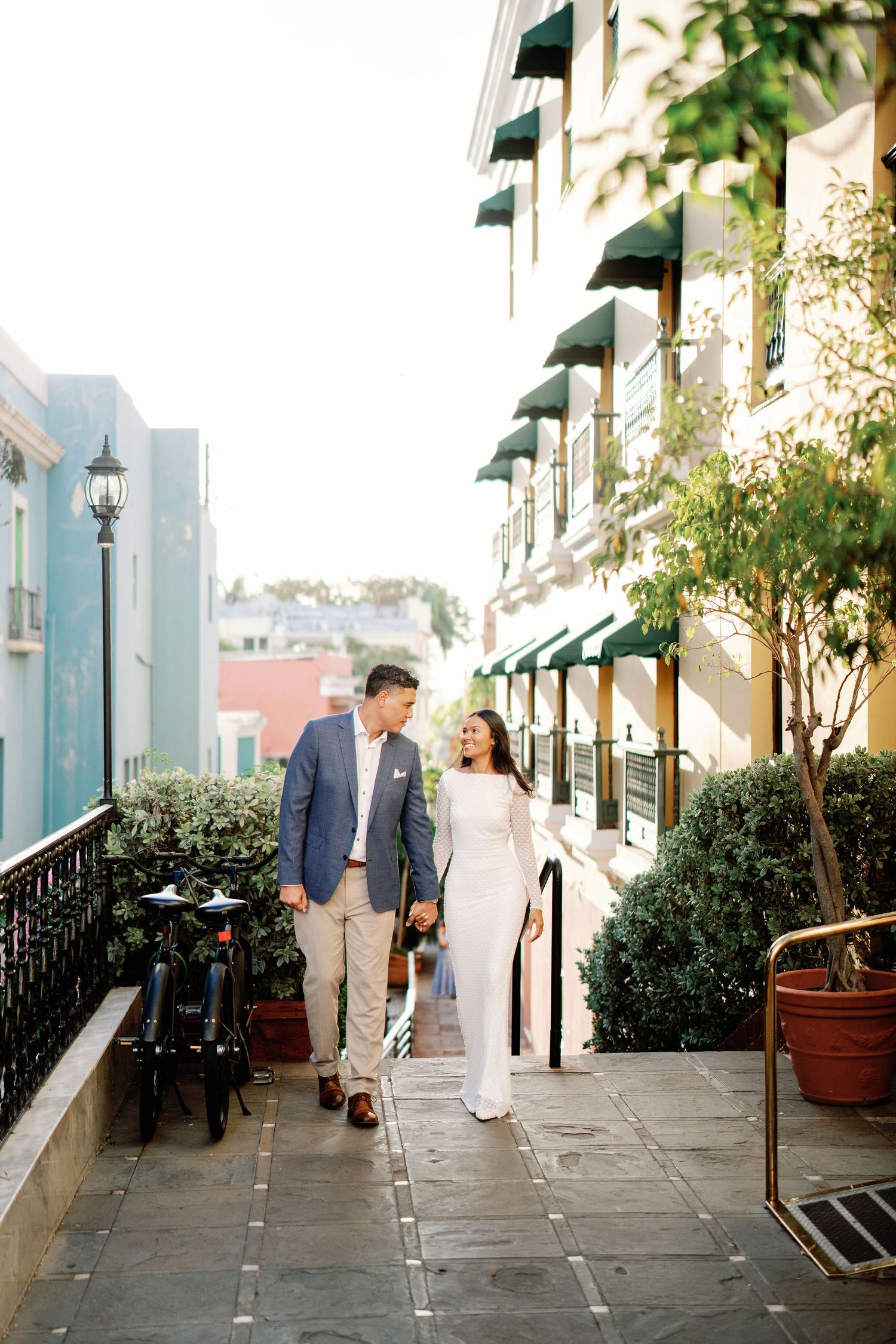 Bride in white gown and groom in blue blazer stroll together along a sunlit building walkway.