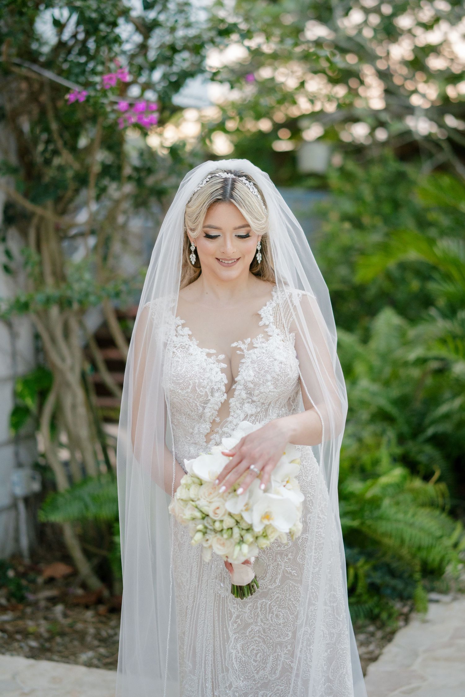 Elegant bride in lace gown and veil holds white bouquet in lush garden setting.