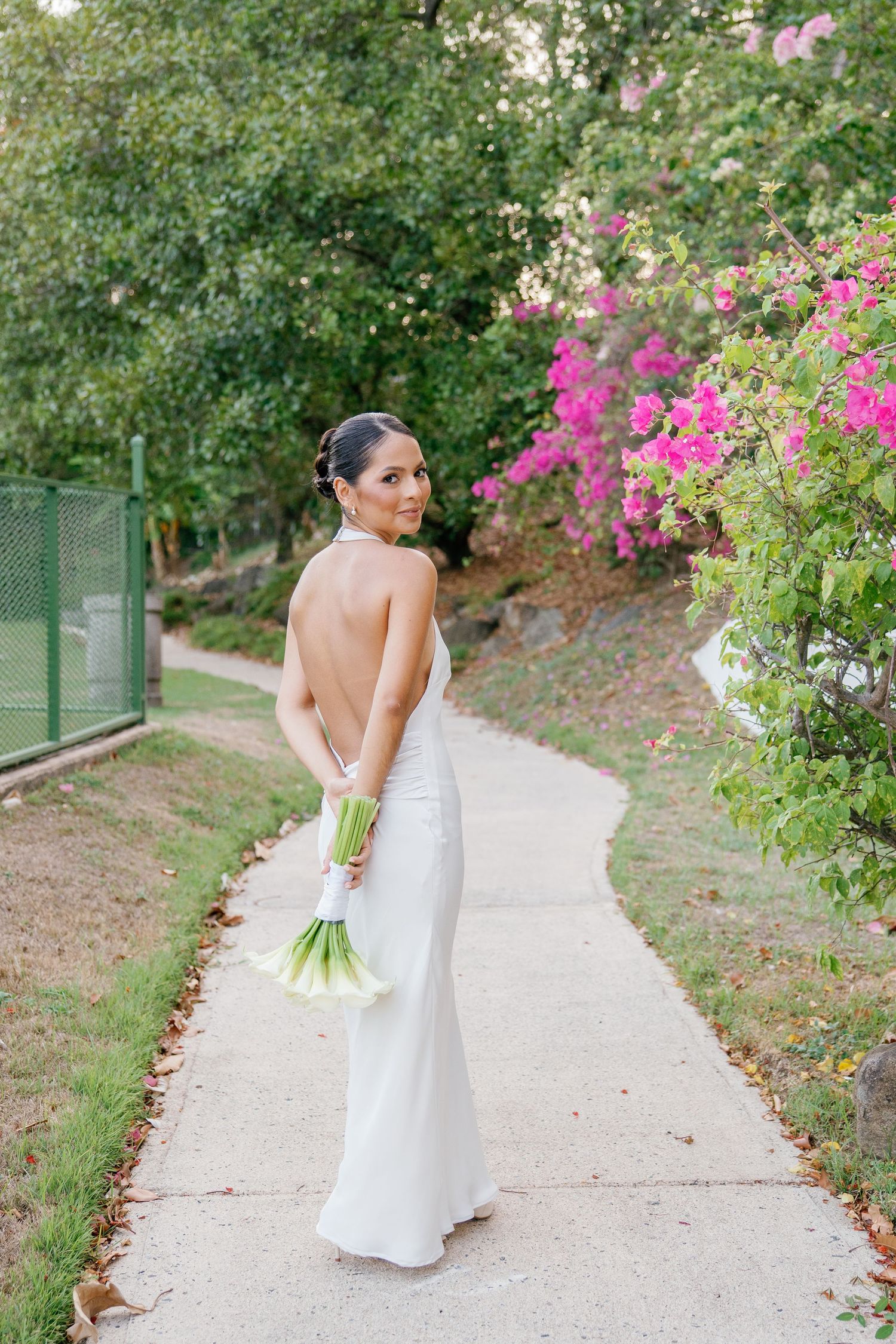 Bride in backless white gown holds green bouquet on garden path with pink bougainvillea blooms.