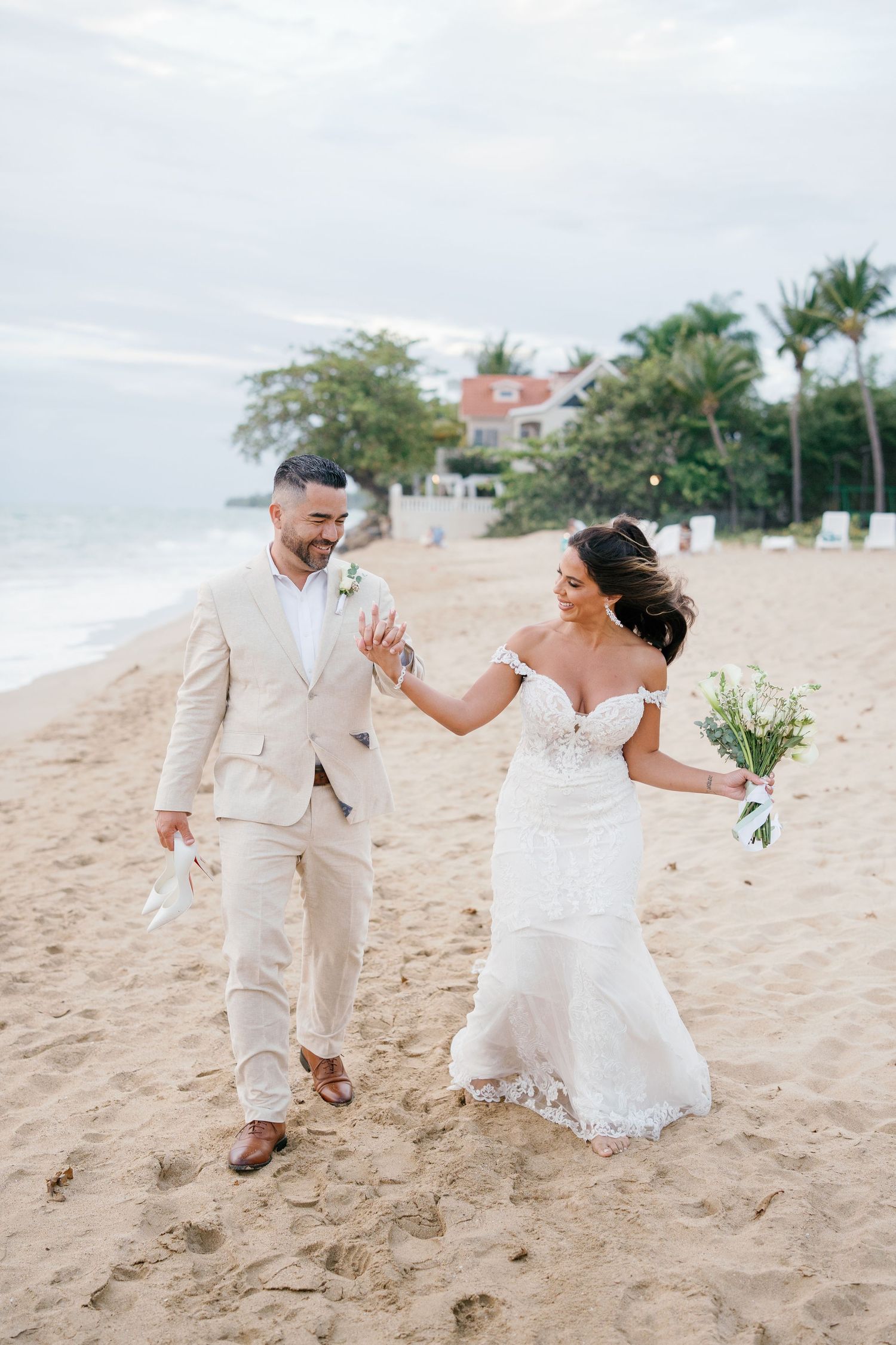 Bride and groom walking hand in hand on a sandy beach, bride in lace mermaid gown holding white bouquet.