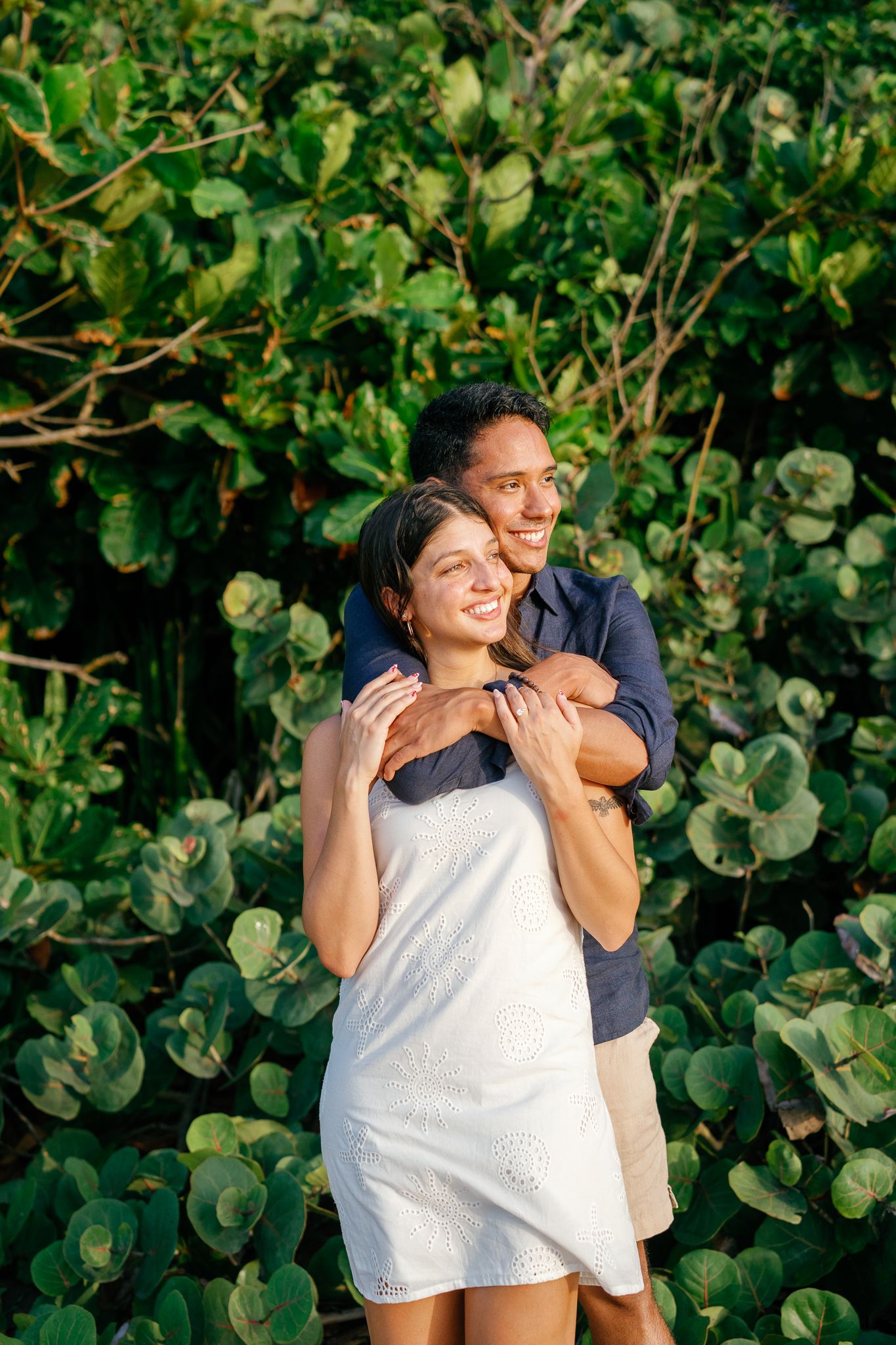 A couple embraces and laughs together against a lush green foliage backdrop in a romantic outdoor setting.