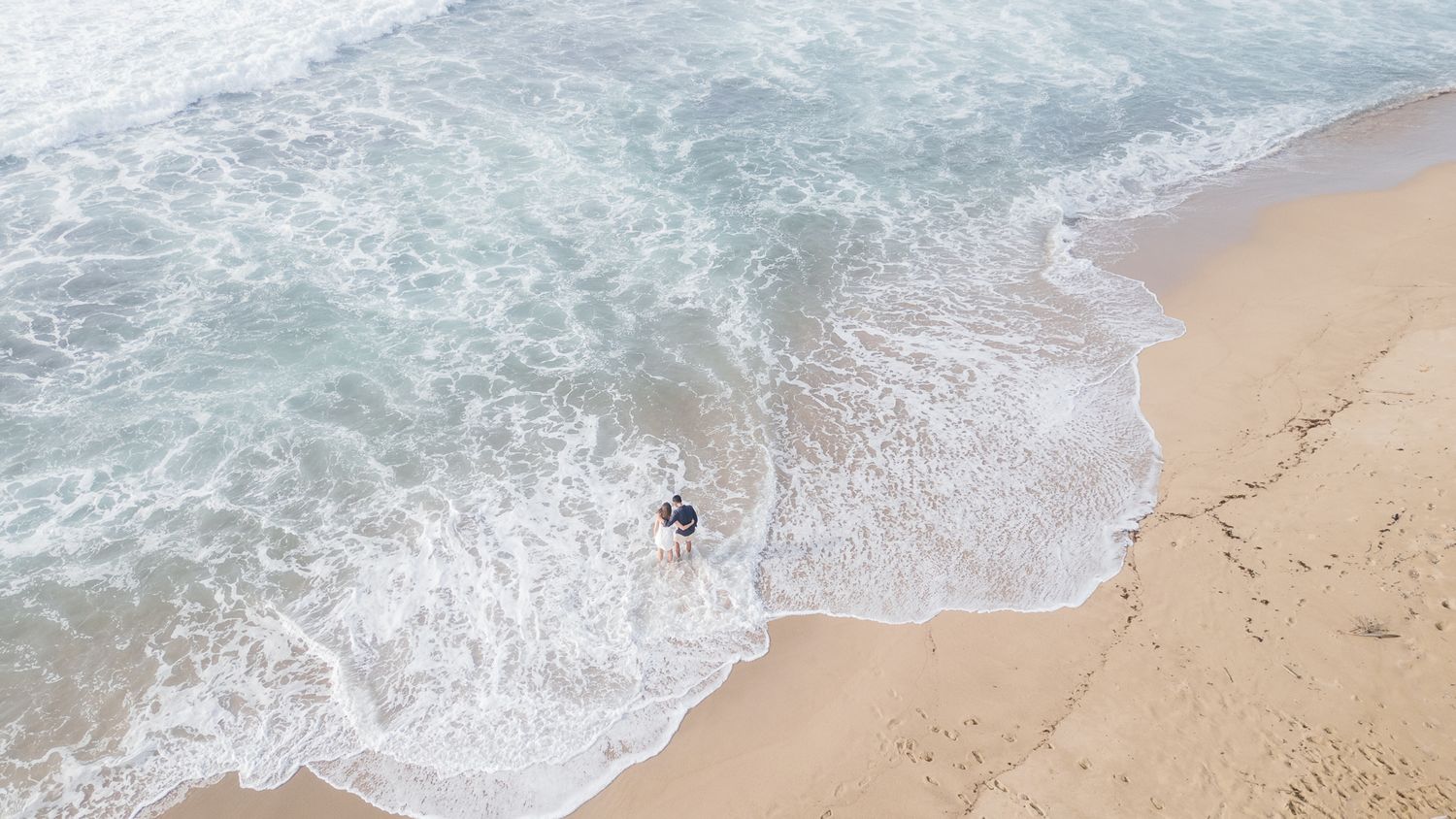 Aerial view of turquoise ocean waves crashing onto a sandy beach with a lone surfer in the water.