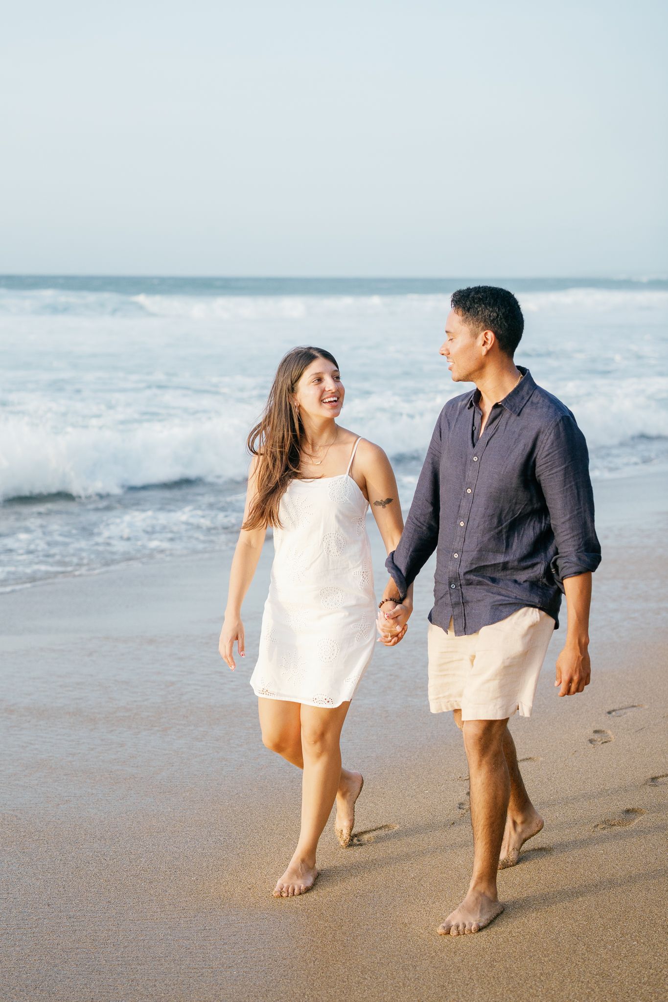 A couple walks hand in hand along a beach at sunset, with ocean waves rolling in the background.