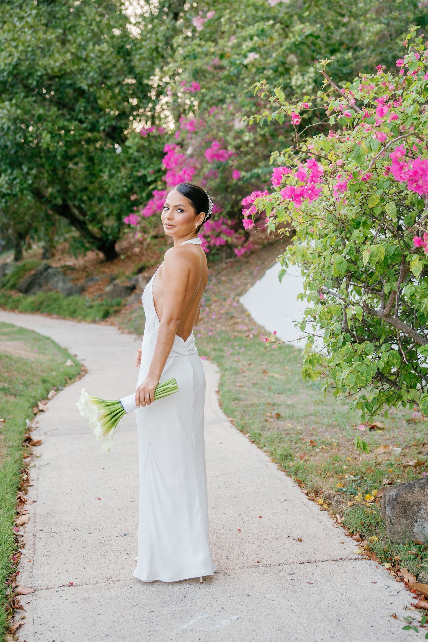 Bride in backless white gown holds white calla lilies on a garden path with vibrant pink bougainvillea blooms.