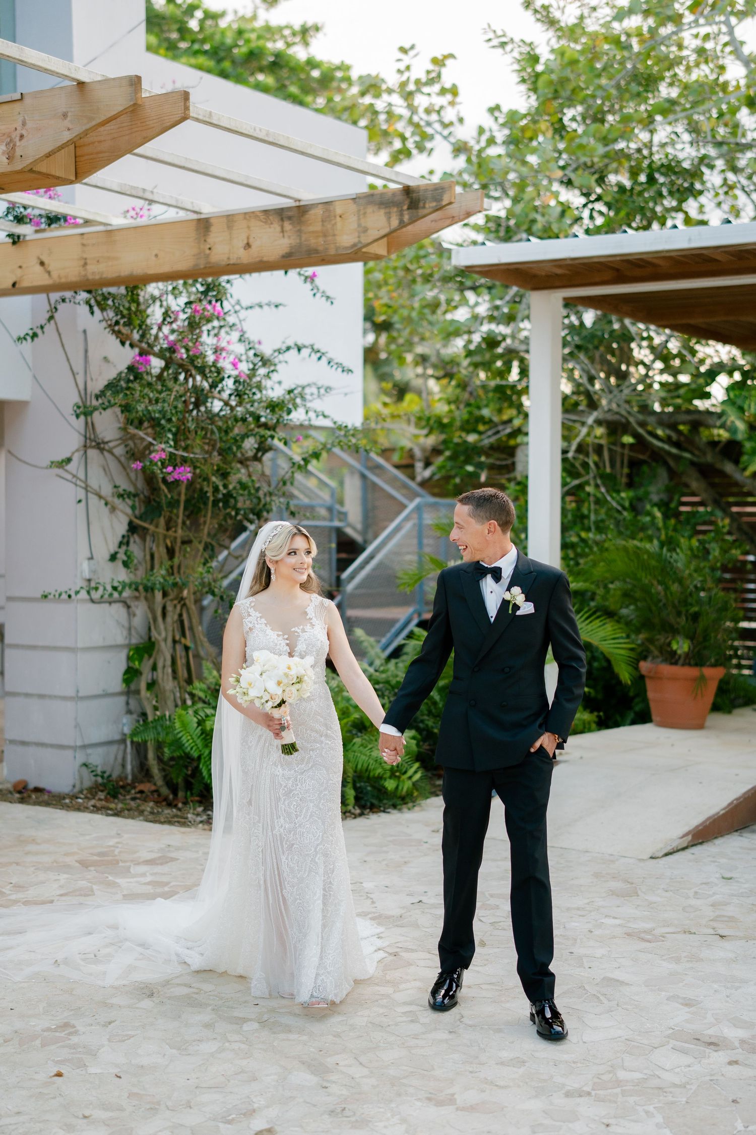 Bride in white gown and groom in black tuxedo hold hands outdoors near tropical greenery and pergola.