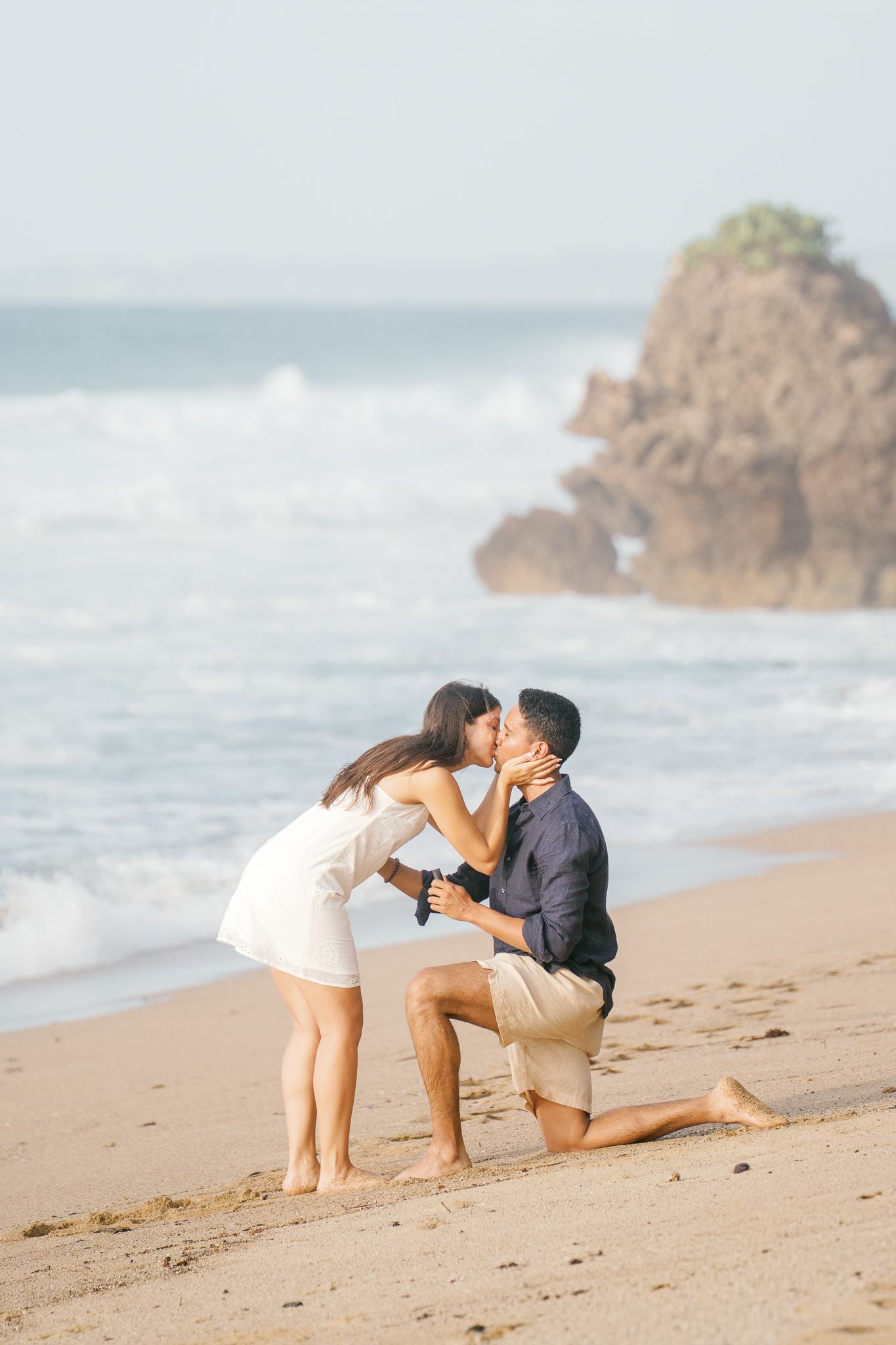 A romantic proposal sequence on a sandy beach with crashing waves and rocky outcrops in the background.