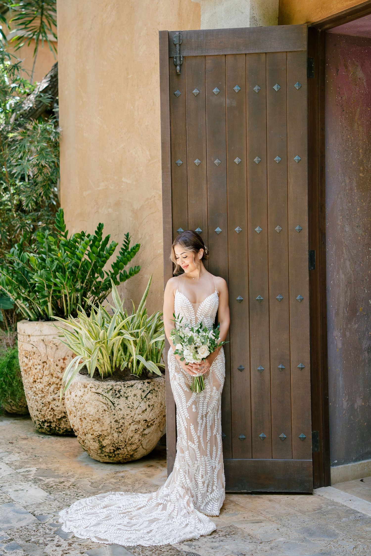Elegant bridal portrait by a rustic wooden door with potted plants and stucco walls in the background.