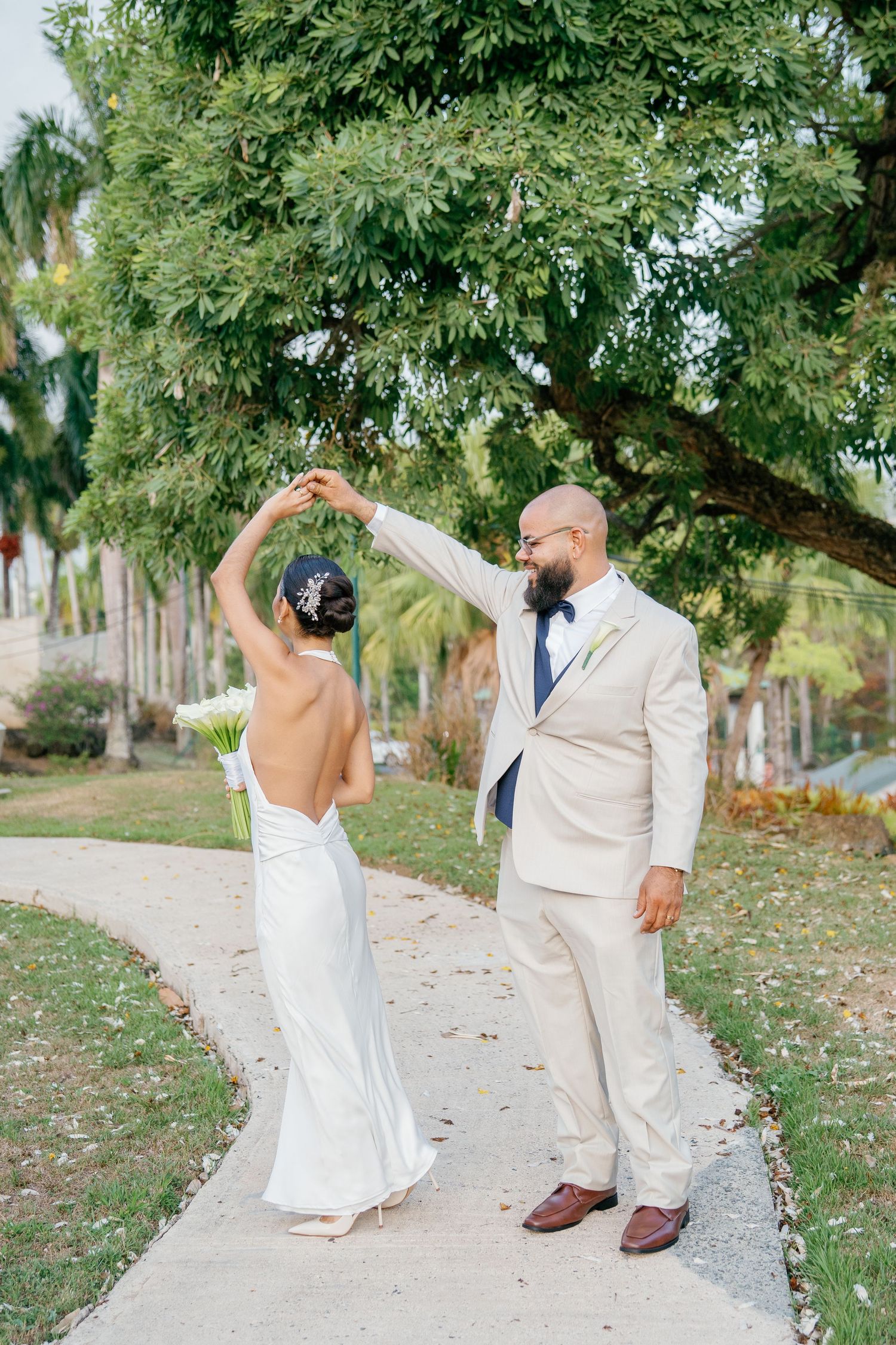 Groom in beige suit twirls bride in backless white wedding gown on garden pathway surrounded by lush tropical trees.