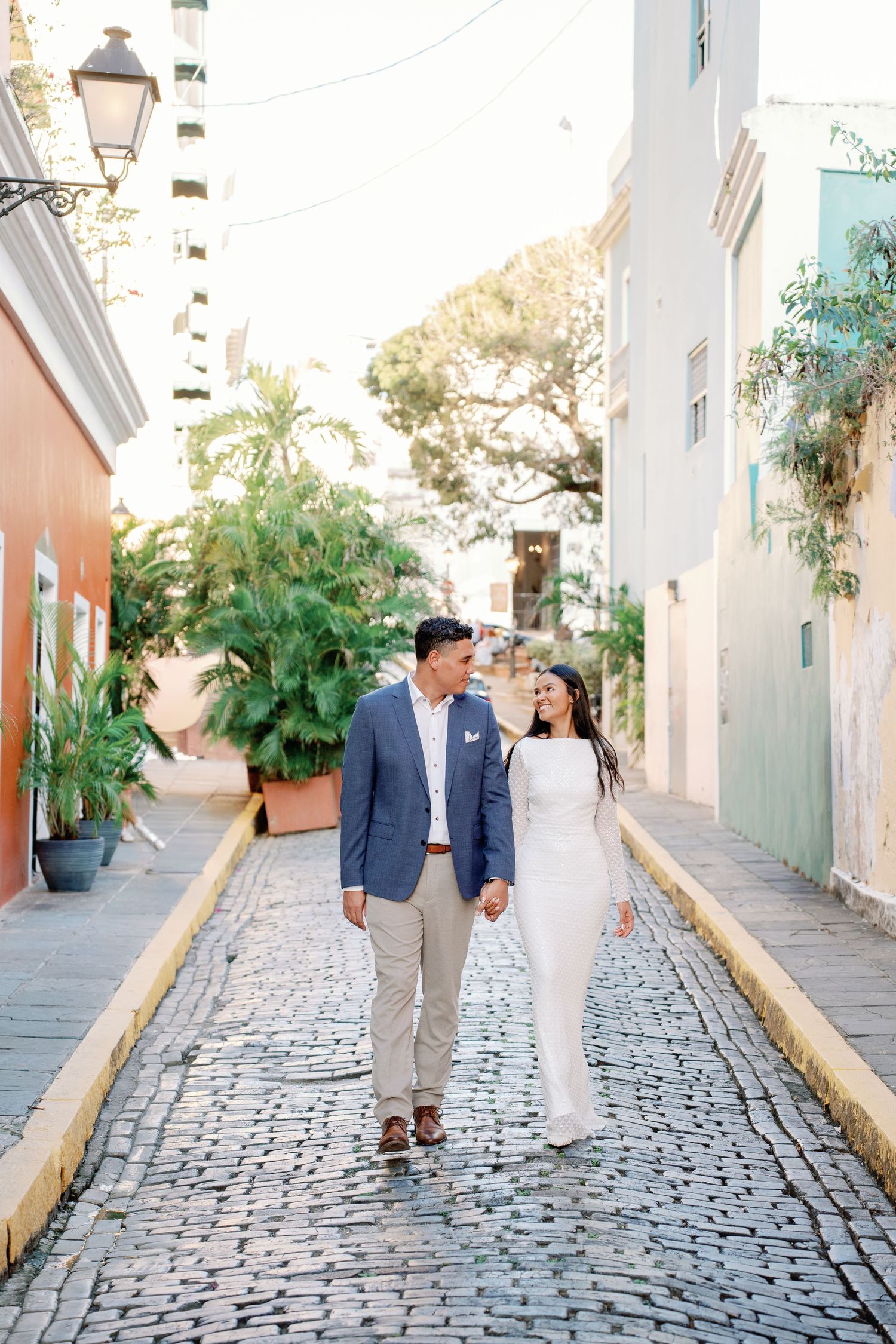 Couple holds hands walking on cobblestone street in Old San Juan, man in blue blazer, woman in white dress.