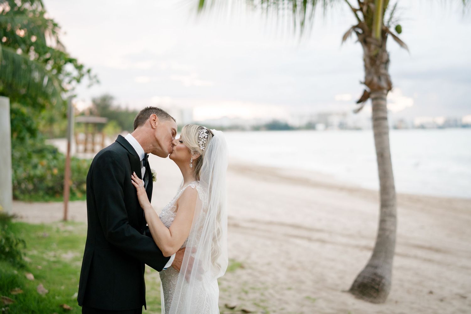 Bride and groom share a romantic beach kiss near a palm tree in wedding attire with ocean views.