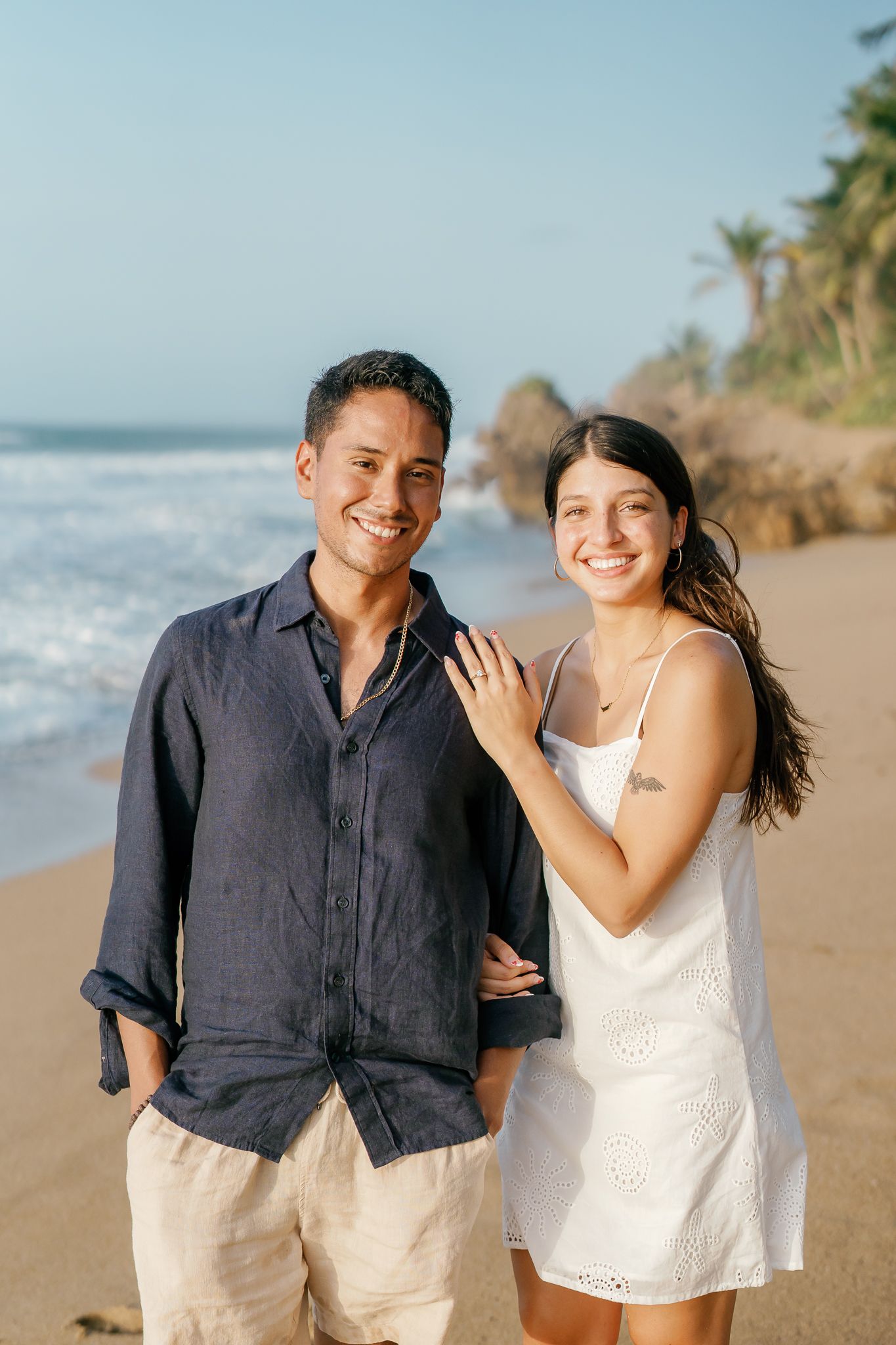 A couple shares romantic moments on a tropical beach with palm trees and ocean waves in the background.