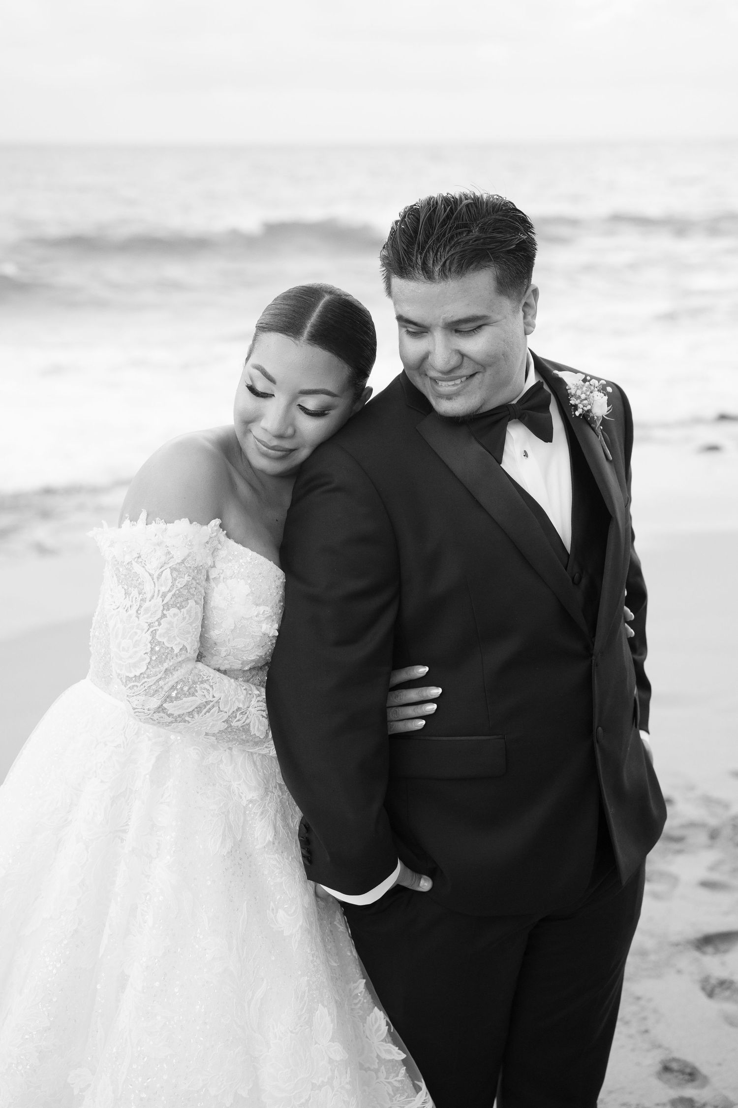 Black and white photo of bride leaning on smiling groom in tuxedo on beach.