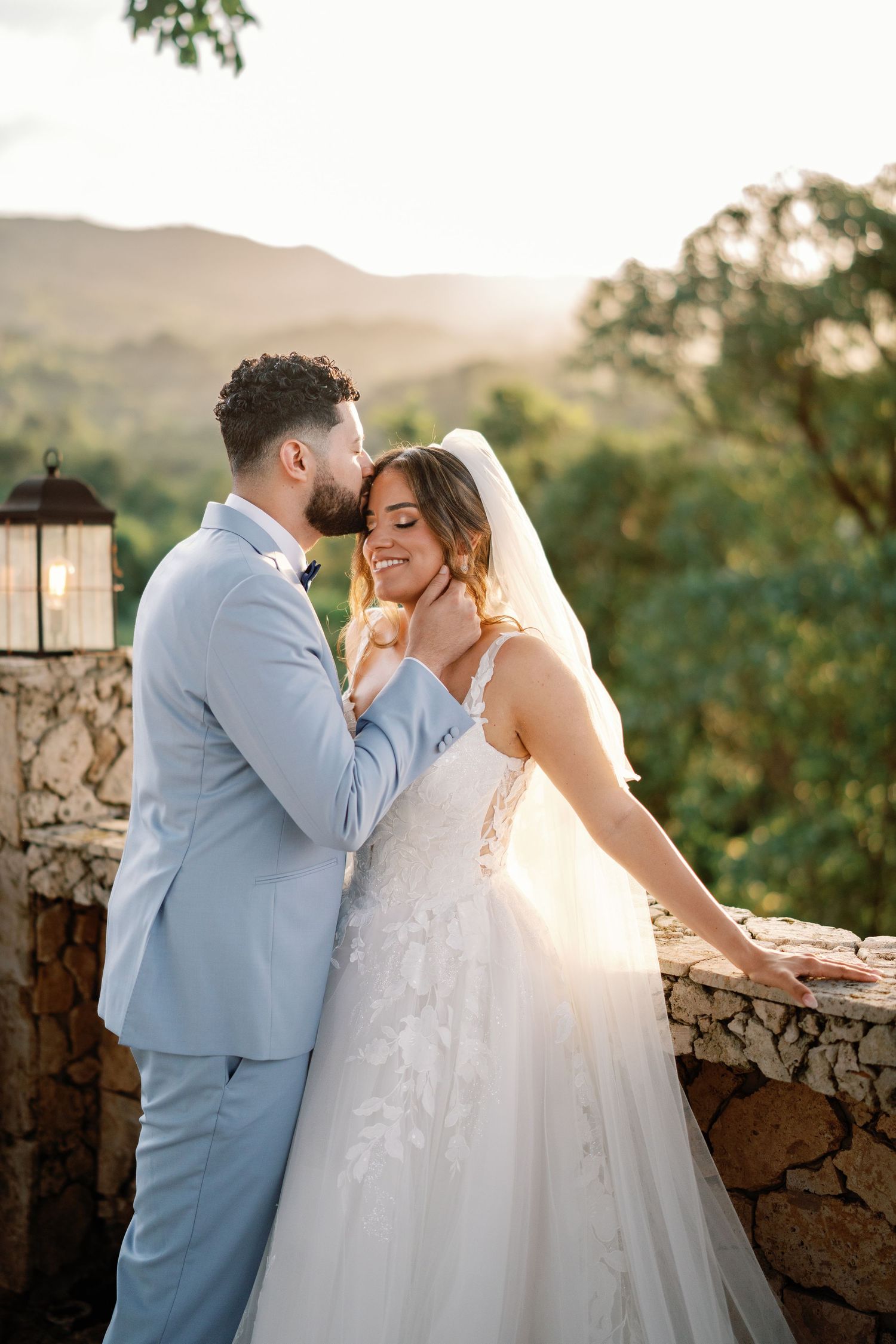Groom in light blue suit kissing bride in white lace gown on stone terrace with mountain views at sunset.