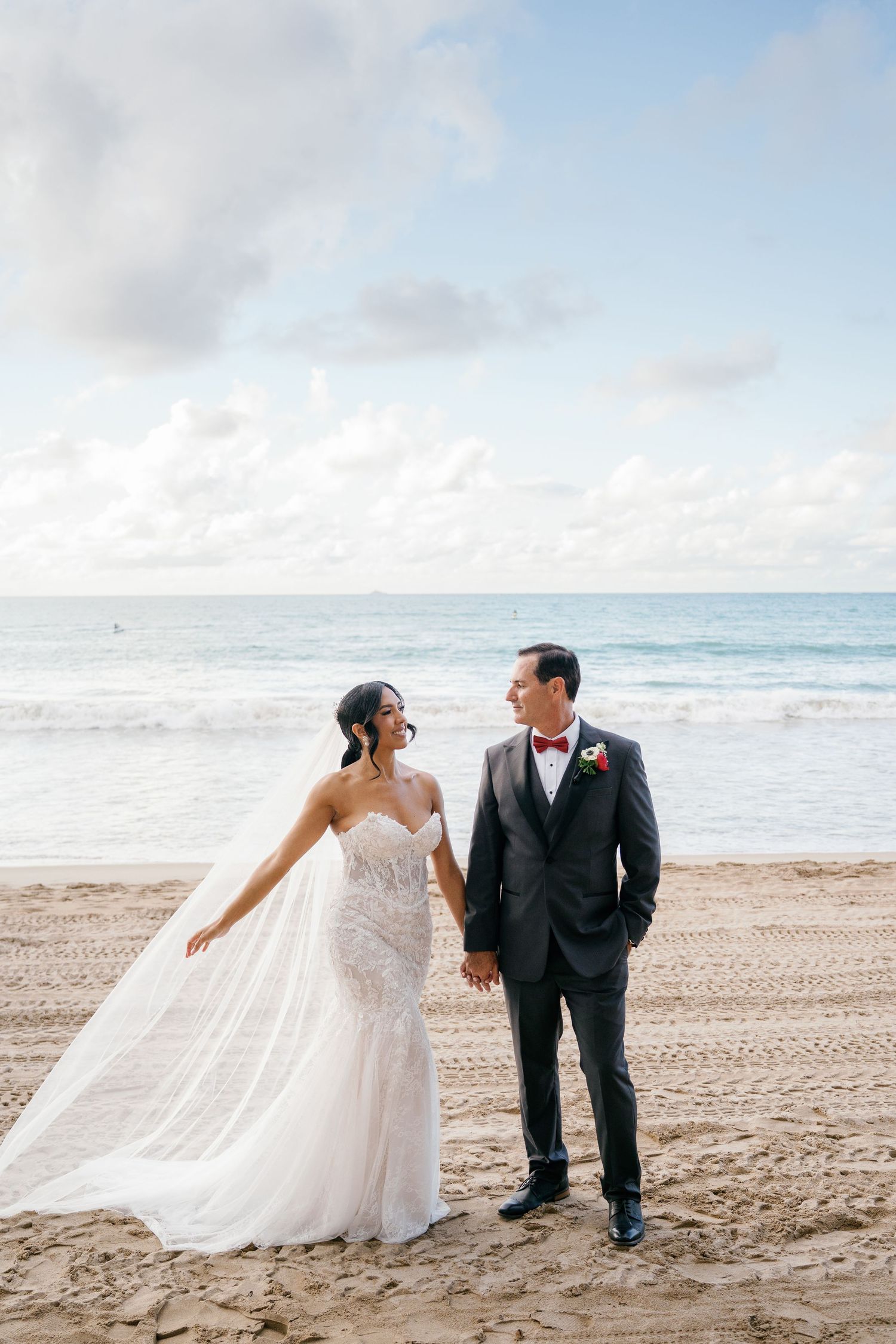 Bride and groom holding hands on a sandy beach, wearing wedding gown and dark suit with red bow tie.