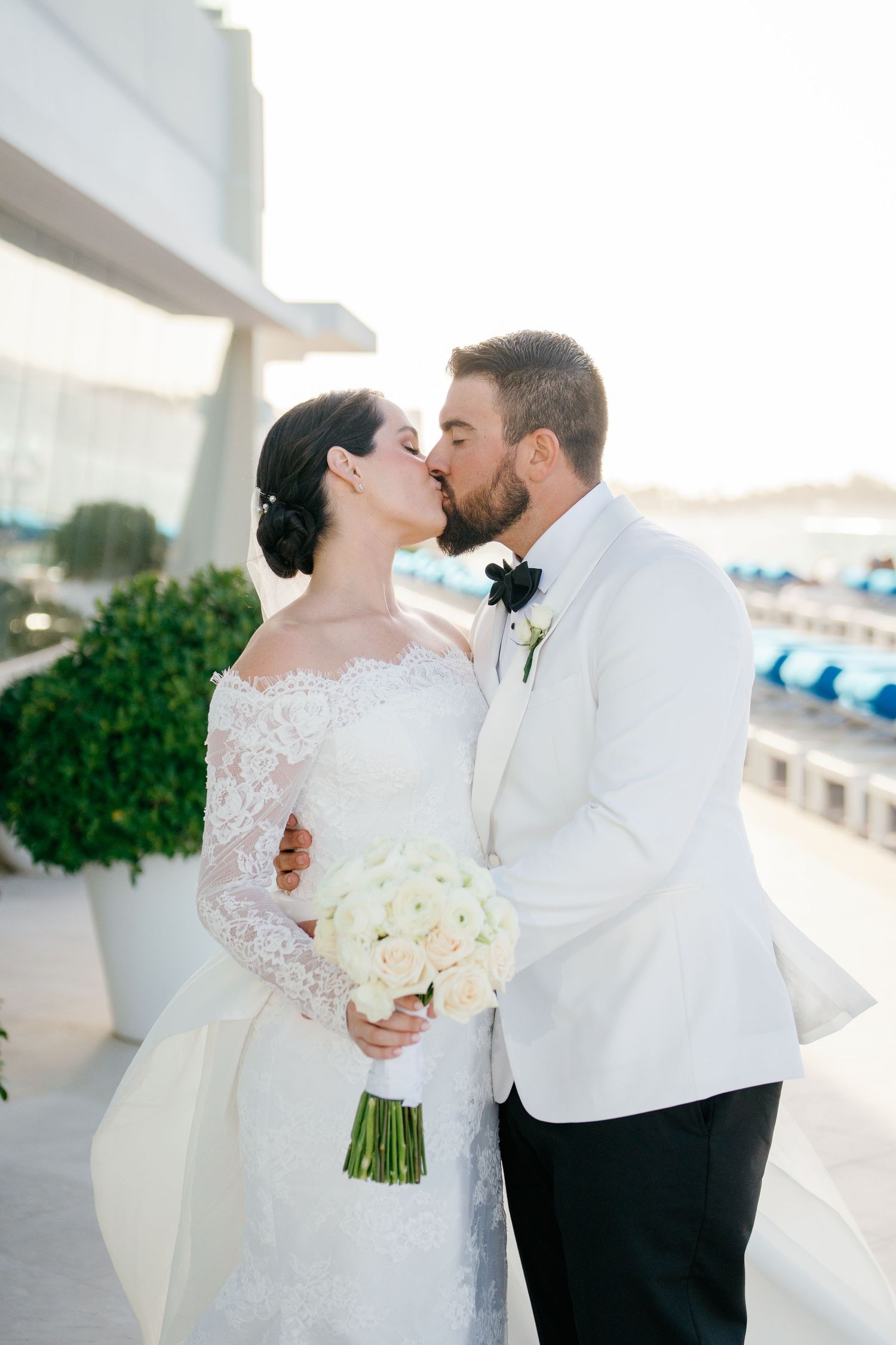 Bride and groom share a romantic kiss, she holds white rose bouquet in lace gown, he wears white tuxedo jacket.