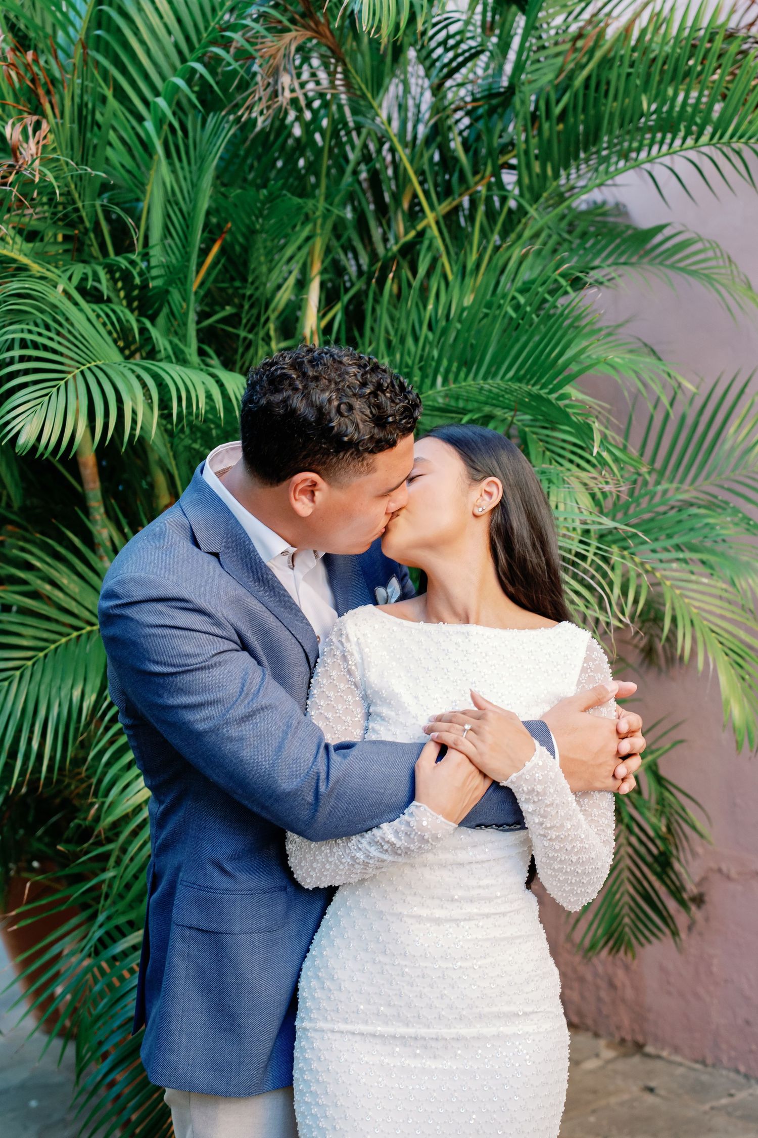 Couple sharing a romantic kiss in wedding attire before lush tropical palm trees against a pink wall.