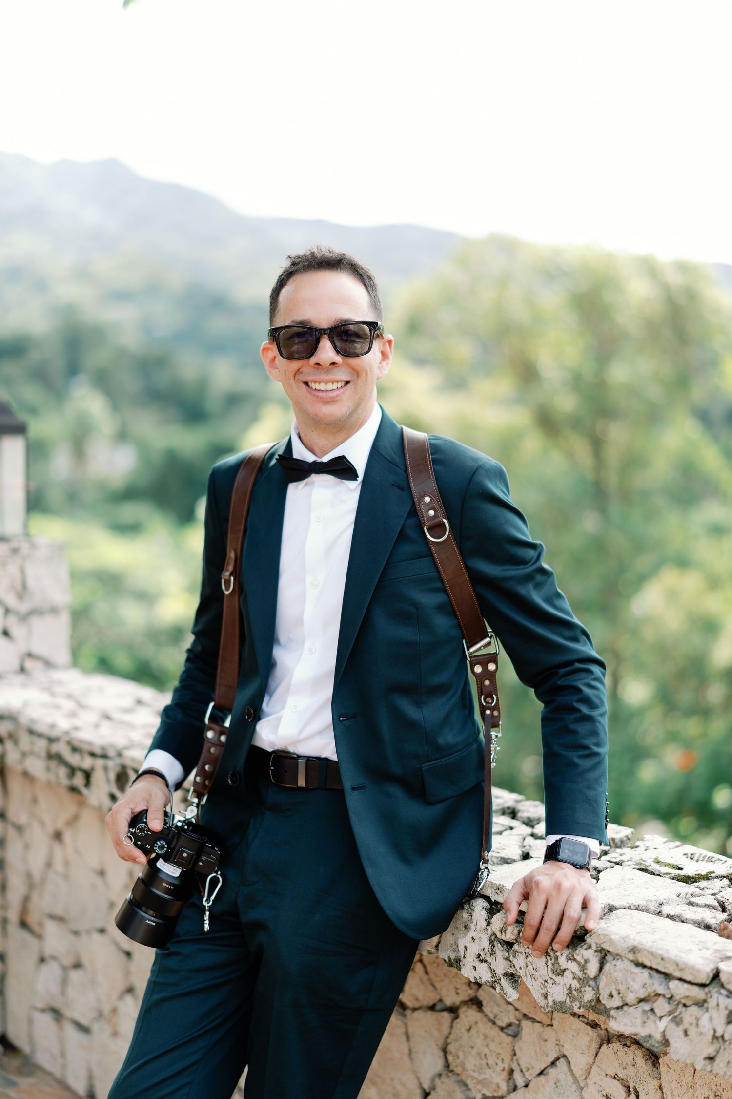 A photographer in a navy suit and white shirt poses against a stone wall with scenic mountain views in the background.