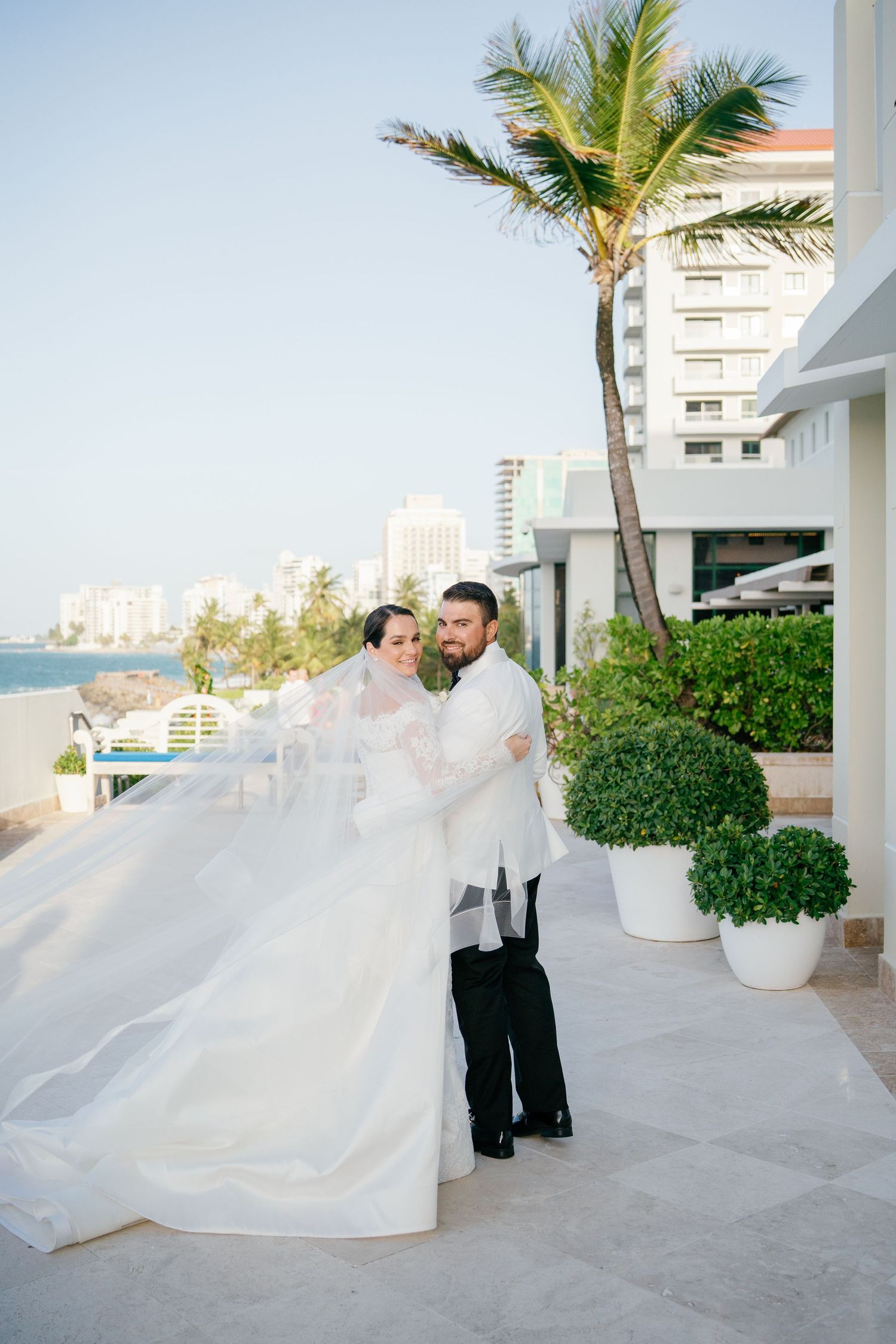 Bride and groom pose on terrace with flowing veil, palm trees and ocean skyline in the background.
