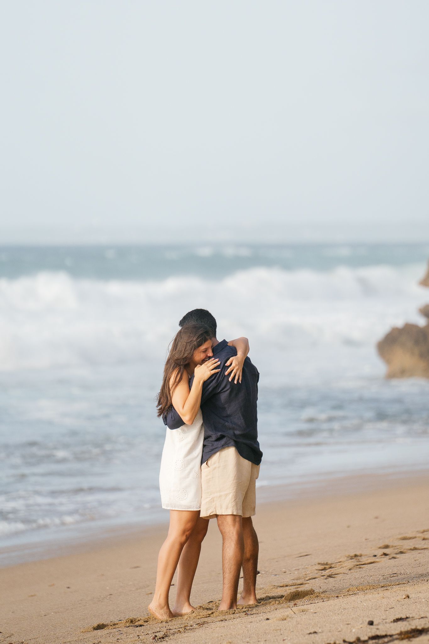 A couple embraces while walking along a sandy beach with ocean waves in the background.