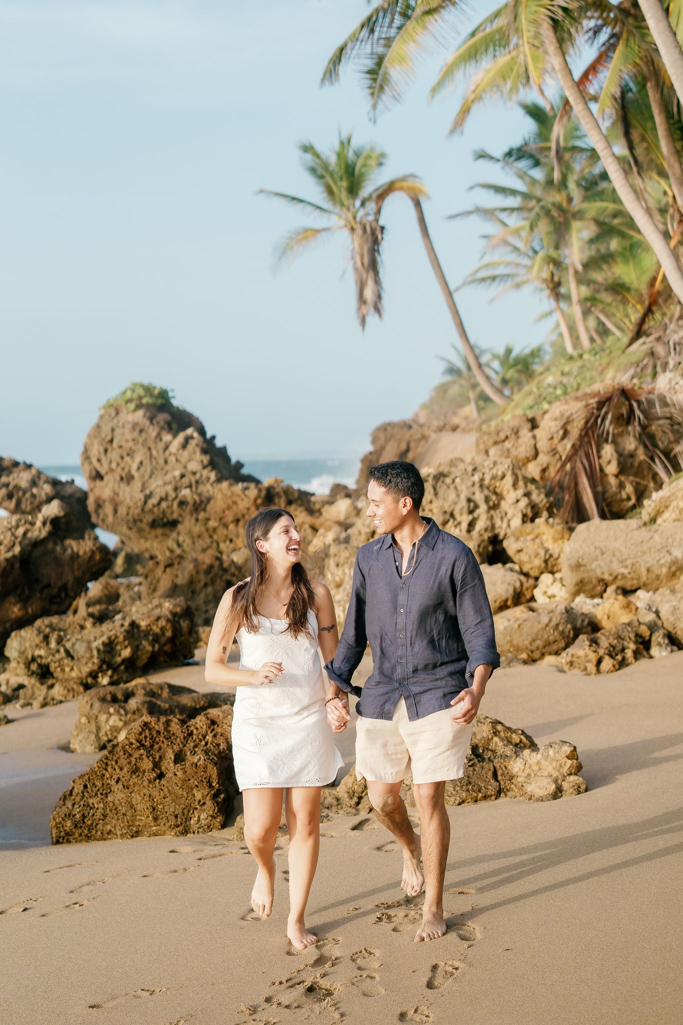 A couple walks together on a tropical beach with rocky formations and palm trees in the background during golden hour.