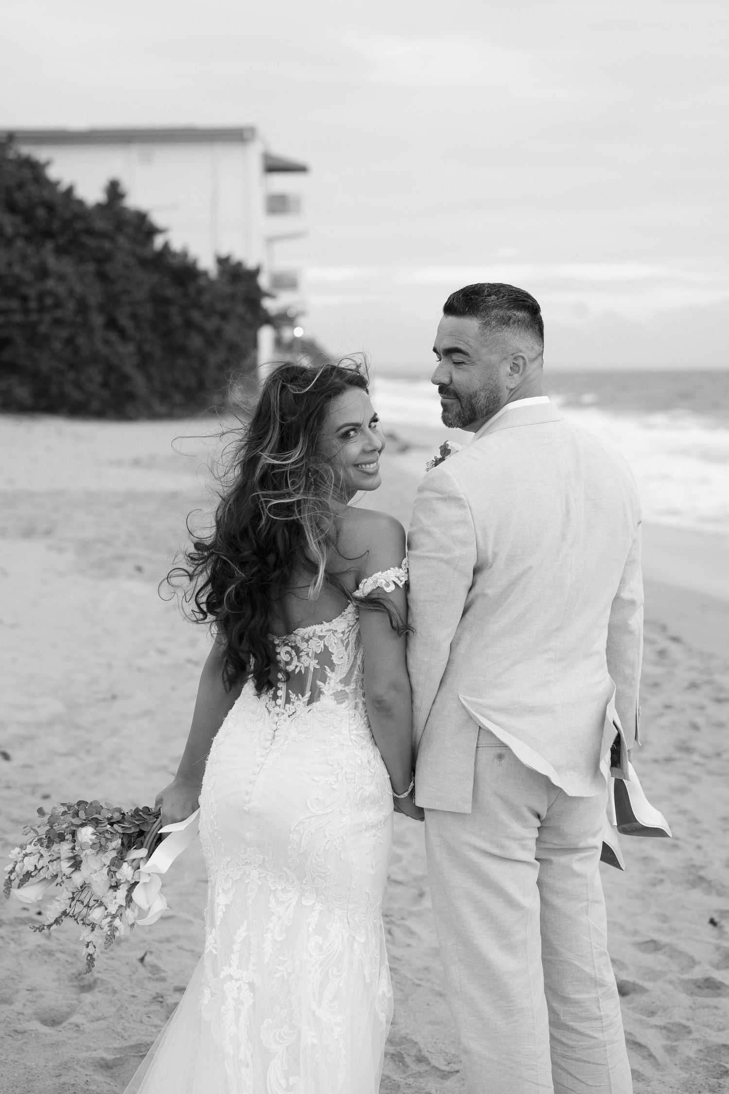 Bride and groom walking on beach, bride in lace gown holding bouquet, groom in light suit.