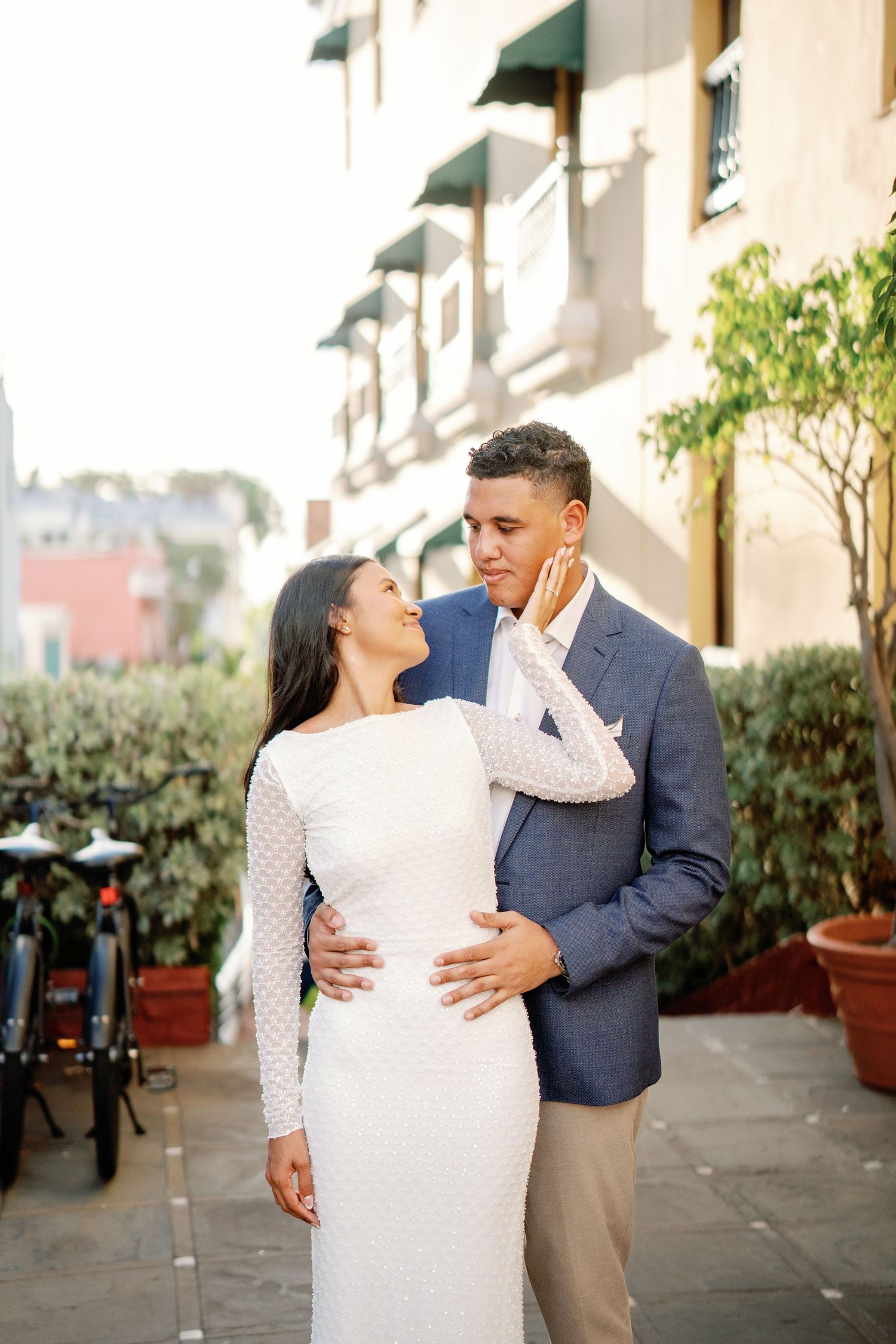 Couple in elegant attire embrace on a sunlit urban street, woman in white gown touches mans face tenderly.