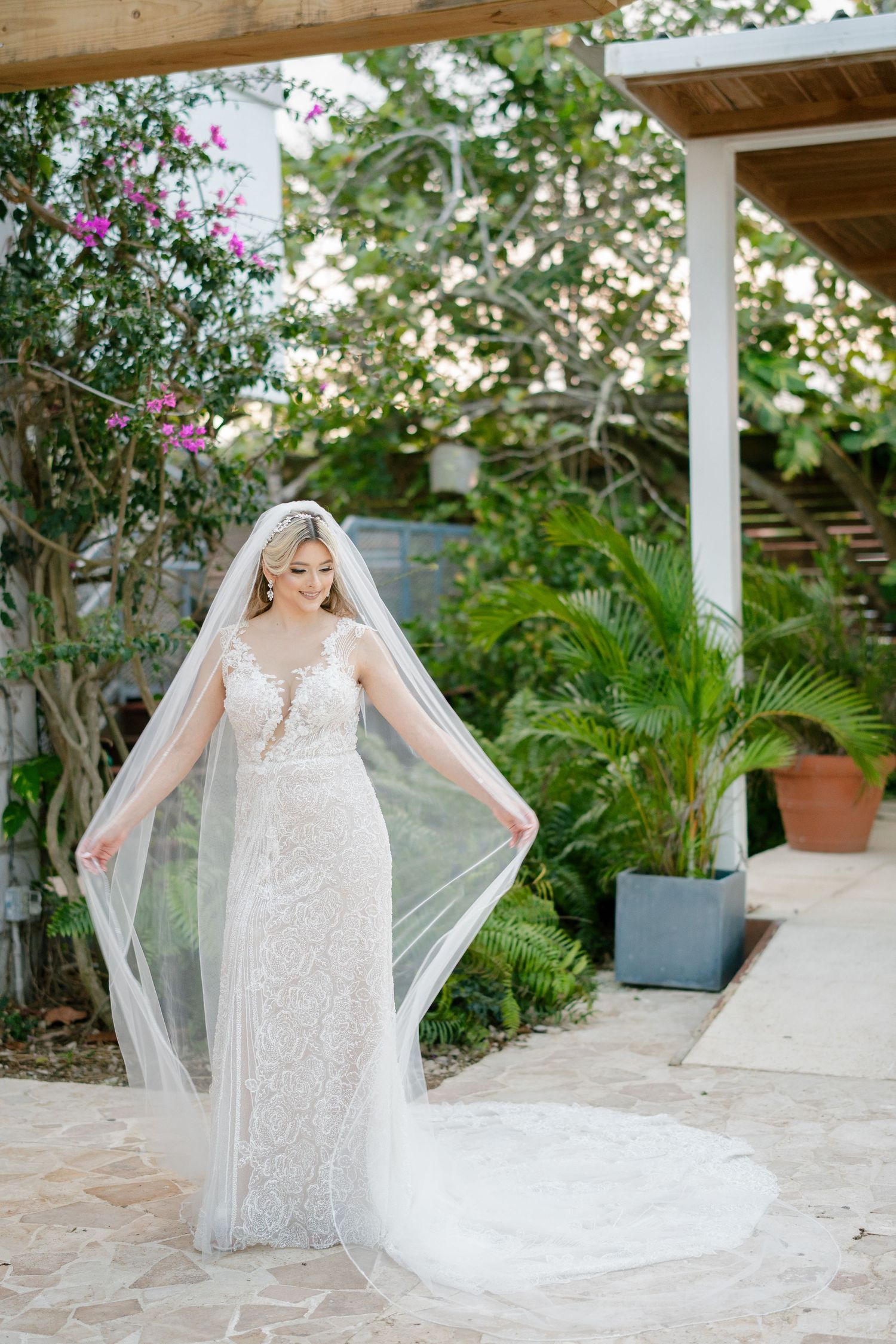 Bride in lace wedding gown and cathedral veil poses gracefully in a lush garden setting with tropical plants.