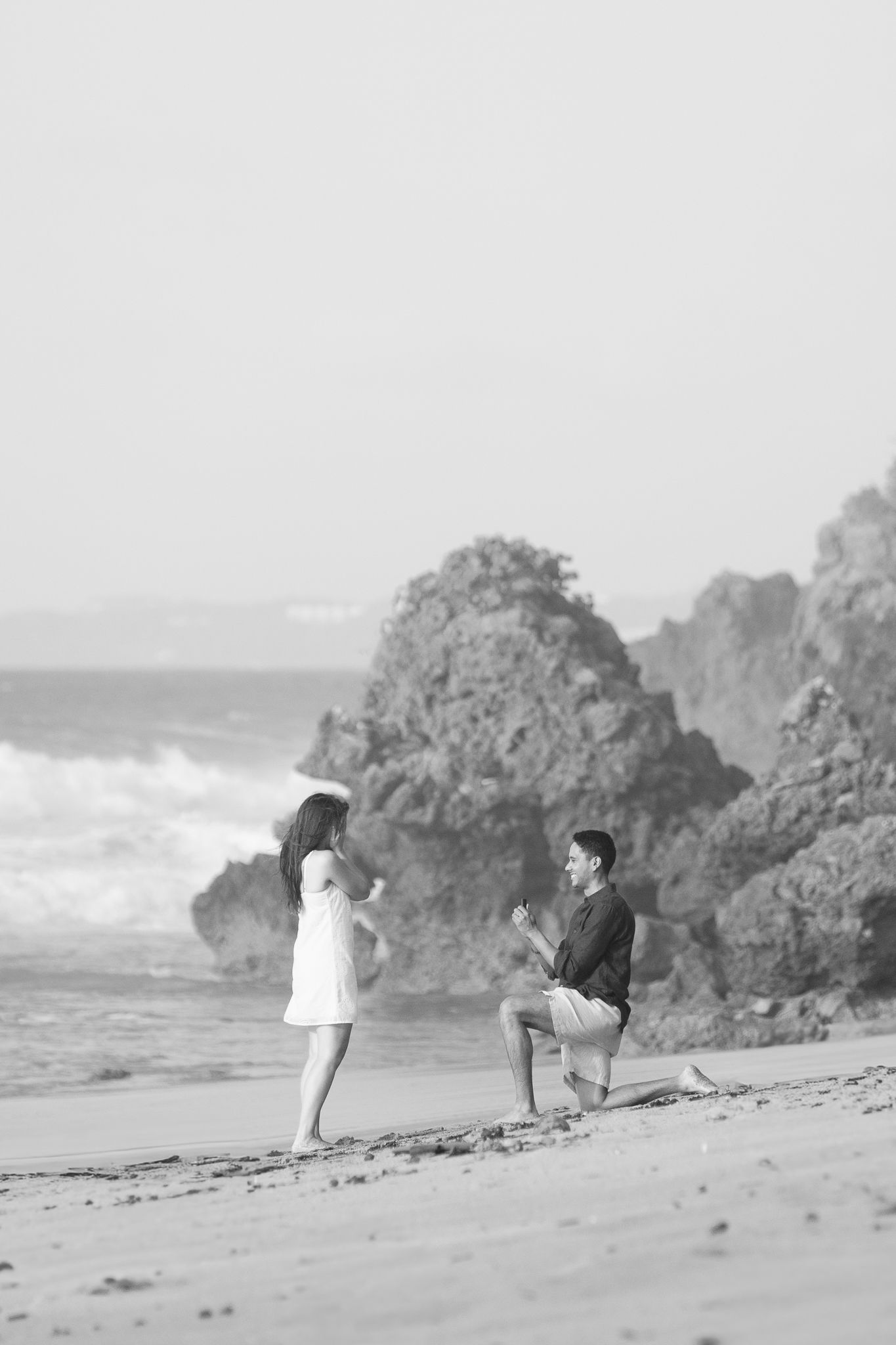 Two people explore a rocky beach shoreline in black and white, bending down to examine the sand near ocean waves.