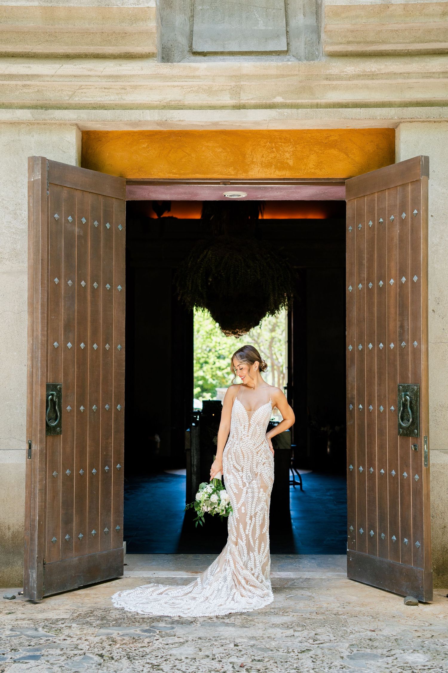 Bride in fitted lace gown poses in grand wooden doorway holding white bouquet.