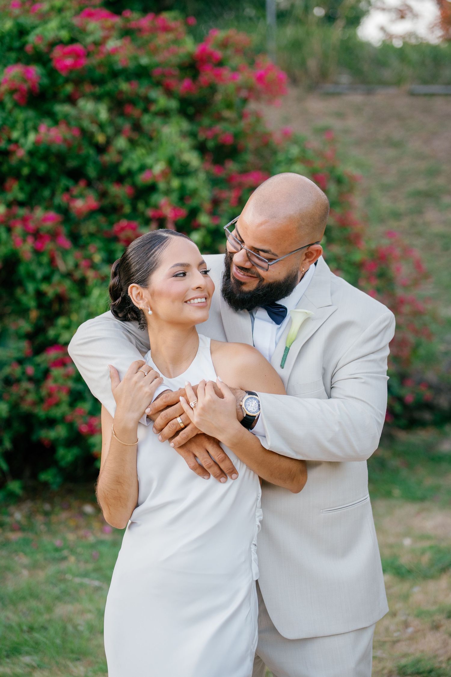 A bride in white dress and groom in beige suit embrace outdoors with pink flowering bushes in background.