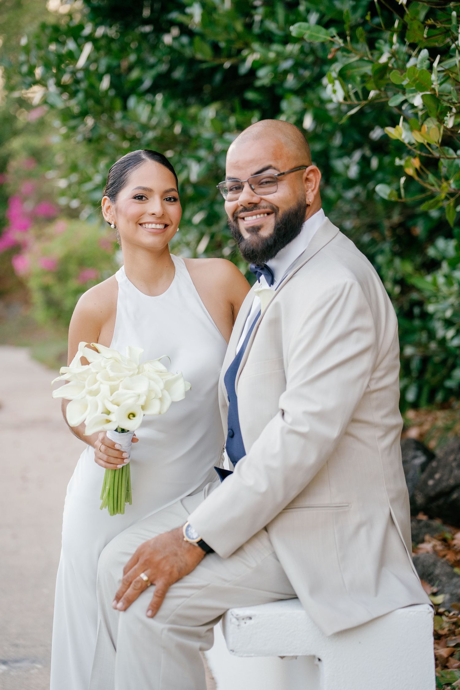 Smiling bride in white halter gown holding calla lily bouquet poses with bearded groom in tan suit outdoors.