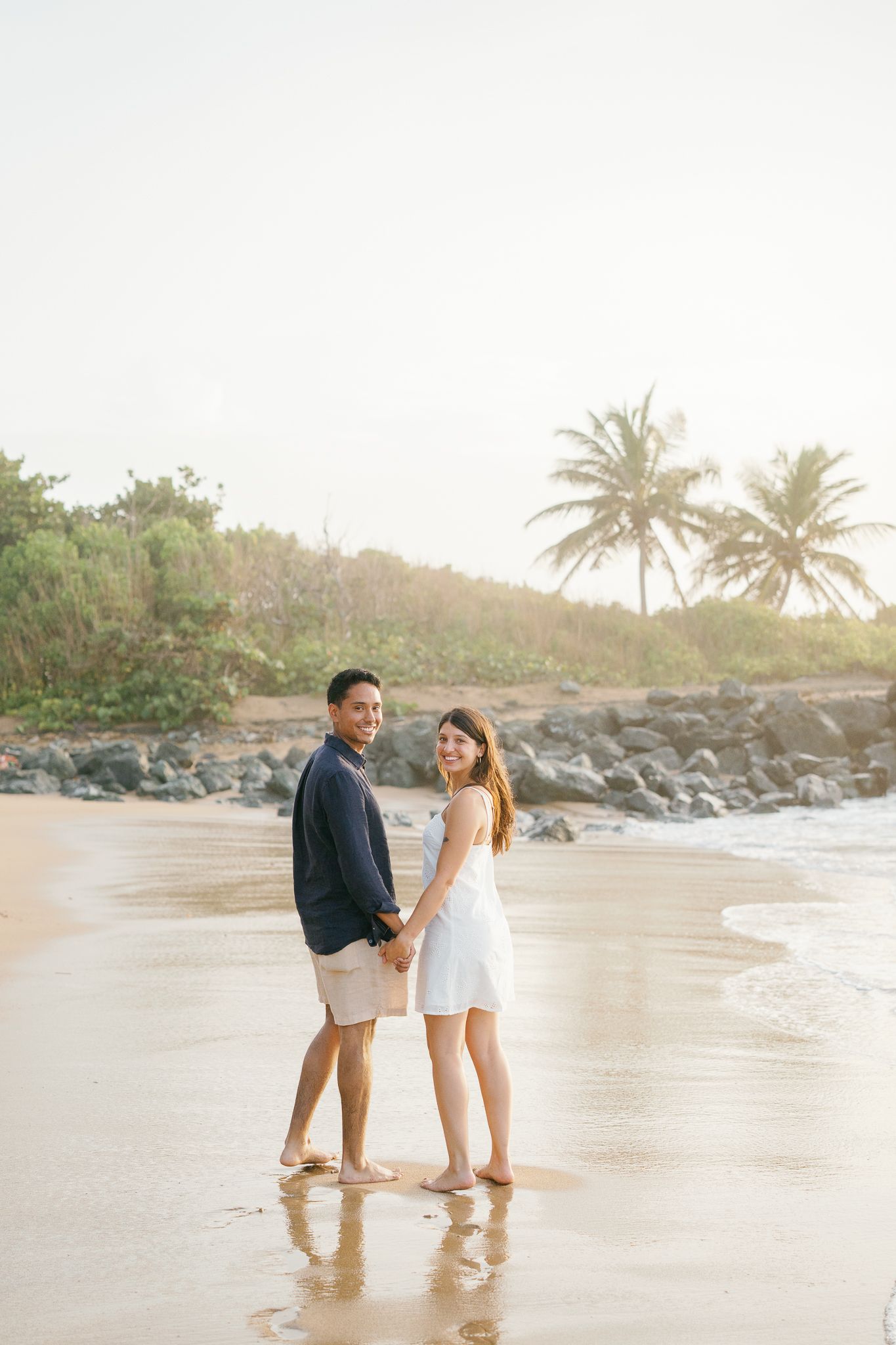 A couple stands together on a tropical beach at sunset with palm trees and rocky coastline in the background.