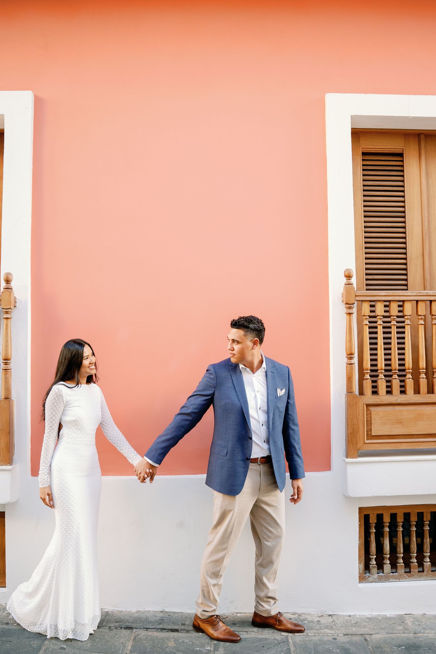 Couple holding hands in front of a coral pink wall, woman in white dress and man in blue blazer.