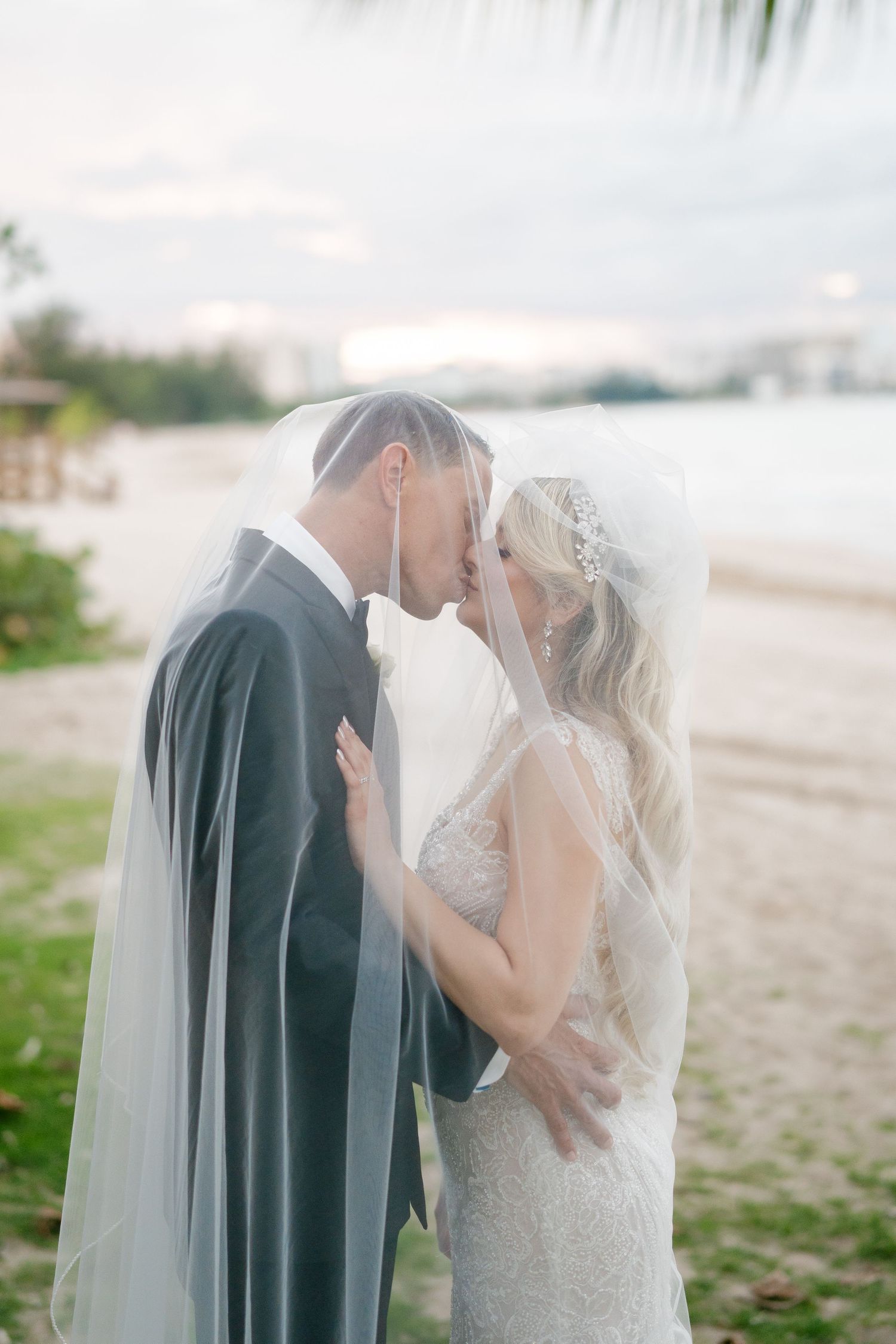 Bride and groom sharing a romantic kiss under a flowing veil on a sandy beach at sunset.