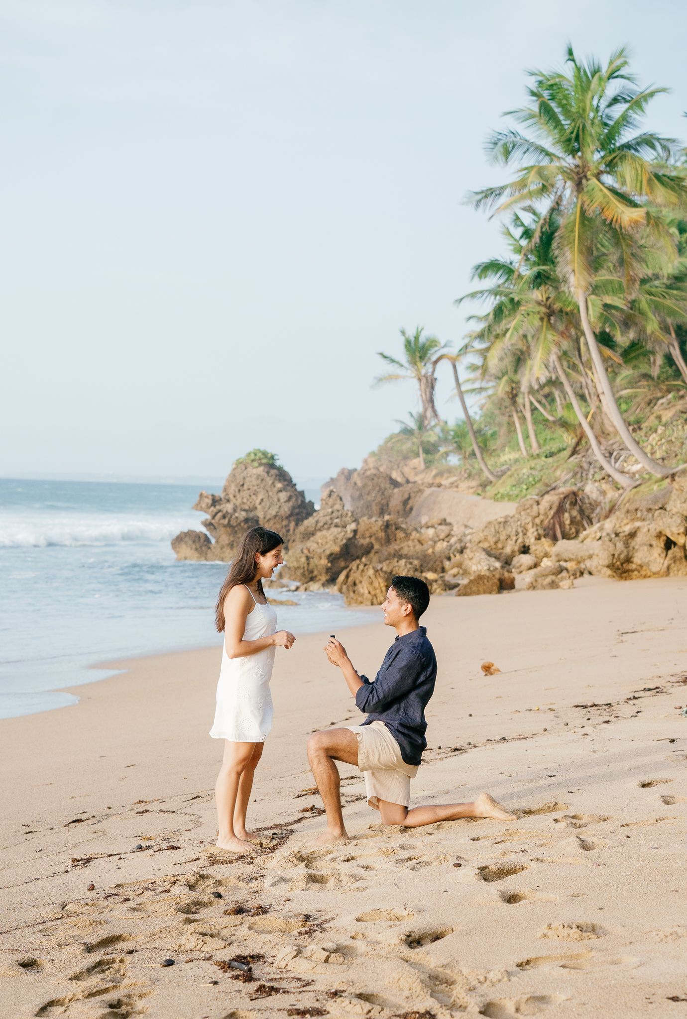 A romantic beach proposal scene unfolds along a tropical shoreline with palm trees and crashing waves in the background.