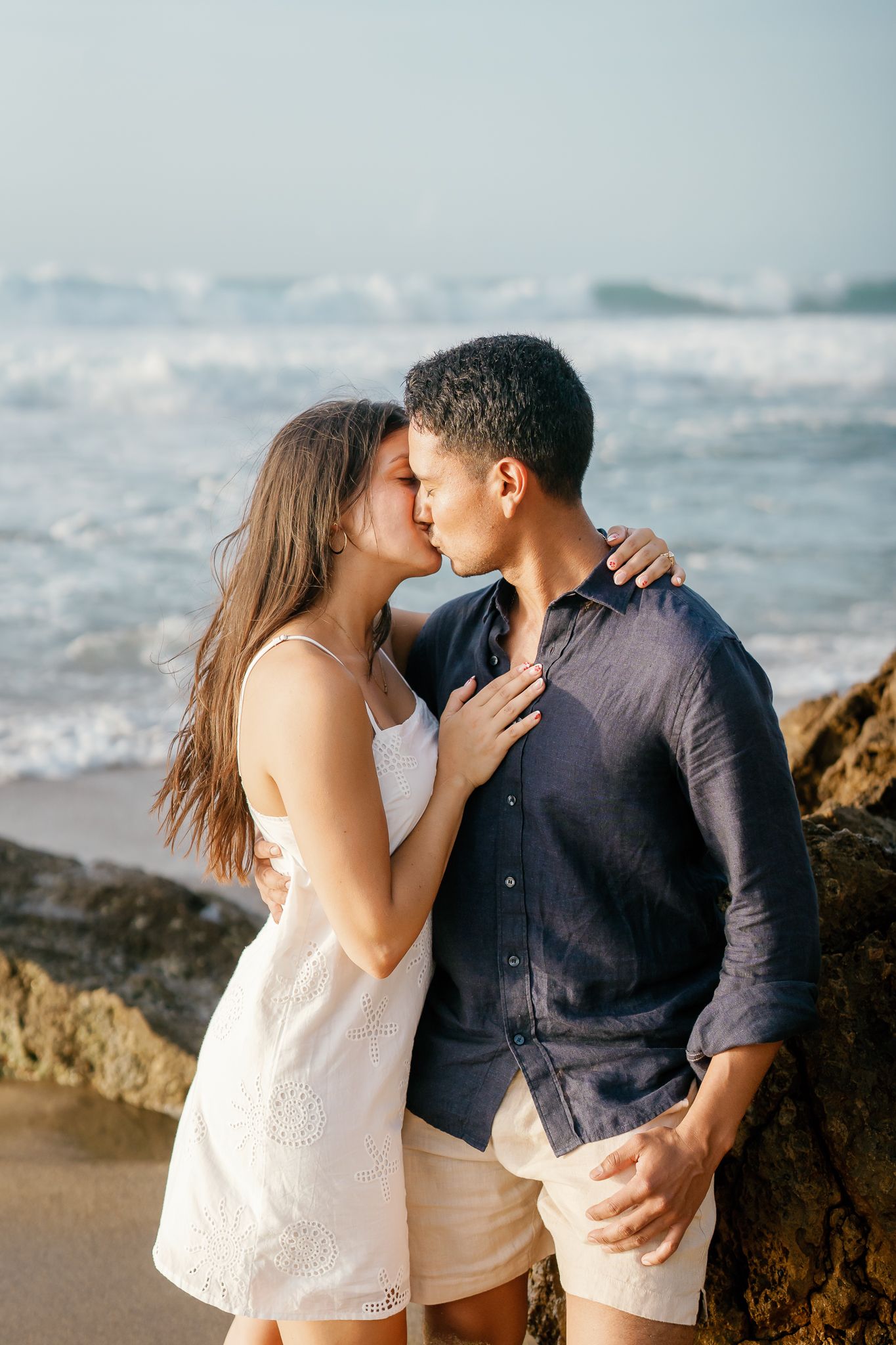 A romantic couple shares a tender kiss on a rocky beach with ocean waves in the background.