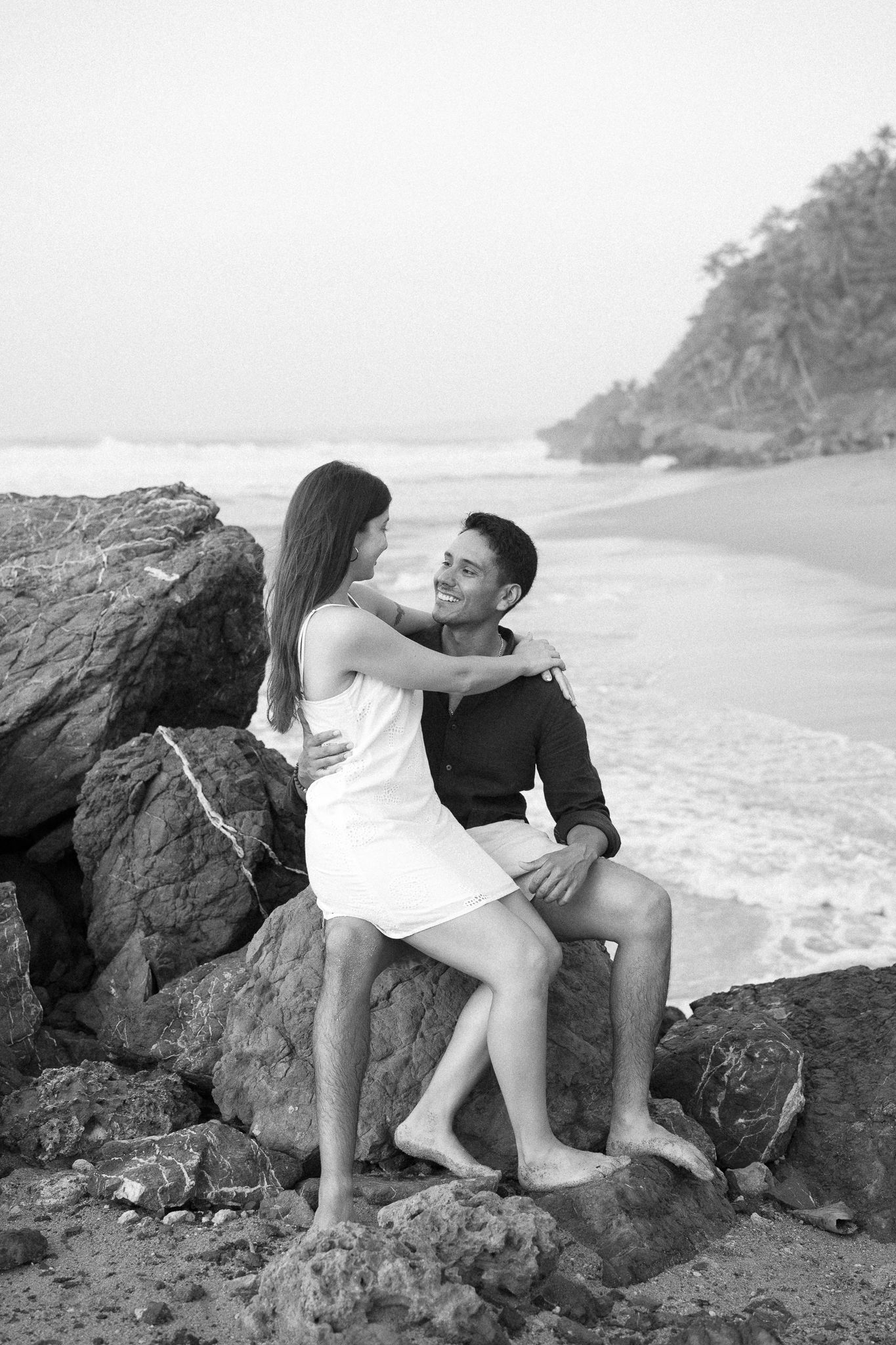 A couple sits intimately together on coastal rocks at the beach with waves and forested cliffs in the background.